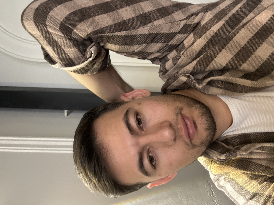 A man with short, dark hair and a trimmed beard poses for a selfie. He wears a white t-shirt layered under a brown and beige plaid flannel. His expression is neutral, with slightly parted lips. The background is a plain wall, and a portion of a white ceiling with a decorative molding is visible in the upper left corner. The image has a slight tilt.