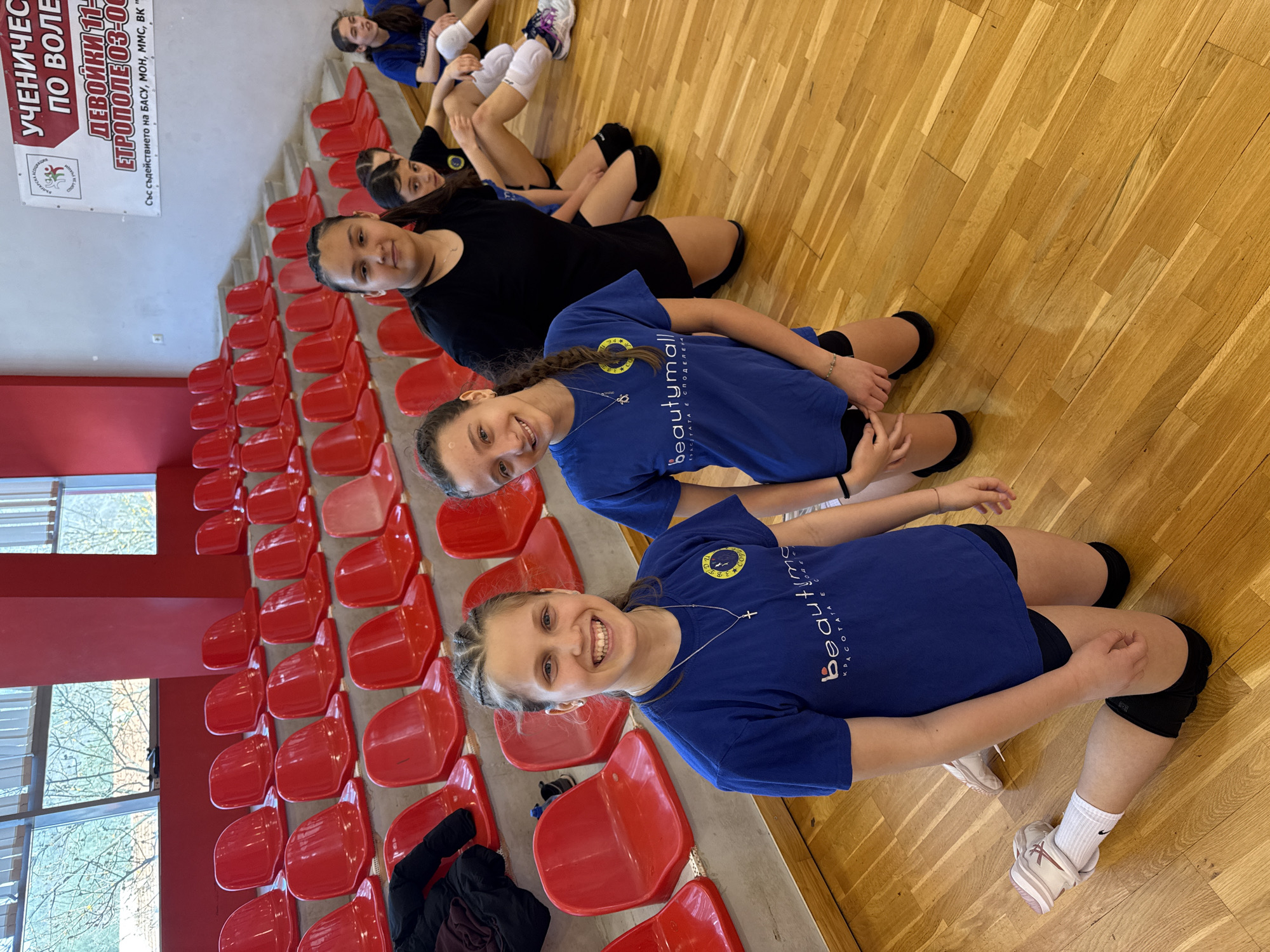 The image shows a group of young female volleyball players posing for a photo in what appears to be a gymnasium or sports hall. The location is Etropole, Bulgaria, as indicated by the sign on the wall. The gymnasium has red bleacher seats. The players are lined up with the two closest to the camera smiling. They are wearing t-shirts of different colors, blue and black. The wooden gymnasium floor is visible. Some players are lying on the floor with others seated. The t-shirts worn by some of the players have the word "beautymall" printed on them. A banner is visible that includes the words "Ученически по волейбол", which translates from Bulgarian to "Schoolchildren's Volleyball".