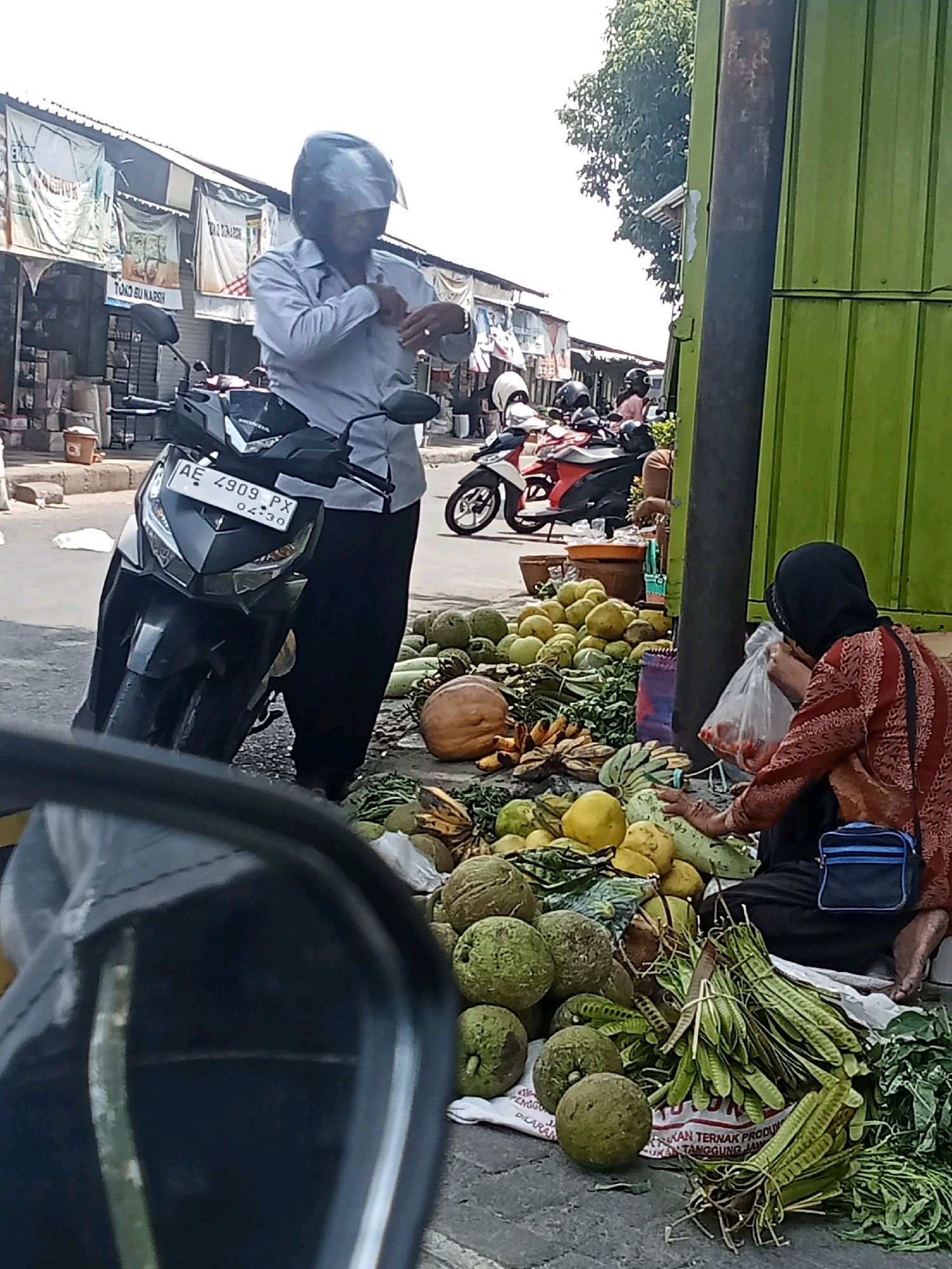 The image depicts a street market scene in Sukowinangun, Indonesia, with a focus on fresh produce. In the foreground, a person wearing a black hijab and an orange and red patterned top sits on the ground, bagging red fruits, likely tomatoes, from a pile of vegetables. Beside her are various fruits and vegetables, including large green gourds, yellow fruits, bananas, and long green beans.
To the left of the produce stall, a motorcycle is parked, and a man in a light blue shirt and black pants, wearing a helmet, stands next to it, appearing to adjust his clothing. The background shows a row of market stalls with white awnings and banners, suggesting a bustling commercial area. Several other motorcycles are visible in the middle ground, indicating transportation common in the area. The overall environment is outdoors, with a paved surface for the market and a green corrugated metal structure visible on the right side of the image. The lighting suggests it is daytime, likely with bright sunlight.