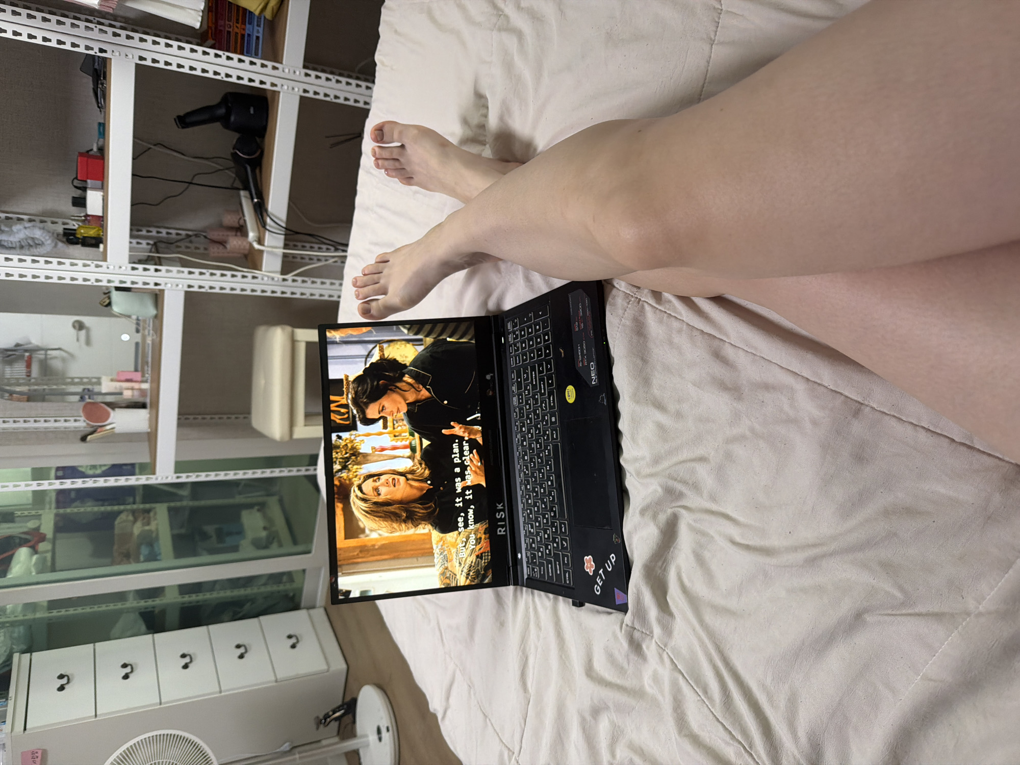 An overhead medium shot captures a person's legs and feet crossed on a bed, positioned in front of an open laptop. The laptop displays a scene from the television show "Friends," featuring actresses Jennifer Aniston and Courteney Cox. The laptop itself is adorned with stickers, including one that reads "GET UP." The bed linens are light-colored and textured. In the background, shelves are visible, holding various items such as cosmetics, hair styling tools, and personal belongings. A white dresser with drawers is also present, along with a floor fan. The scene appears to be within a bedroom, likely in a residential apartment in Dongducheon-si, South Korea.