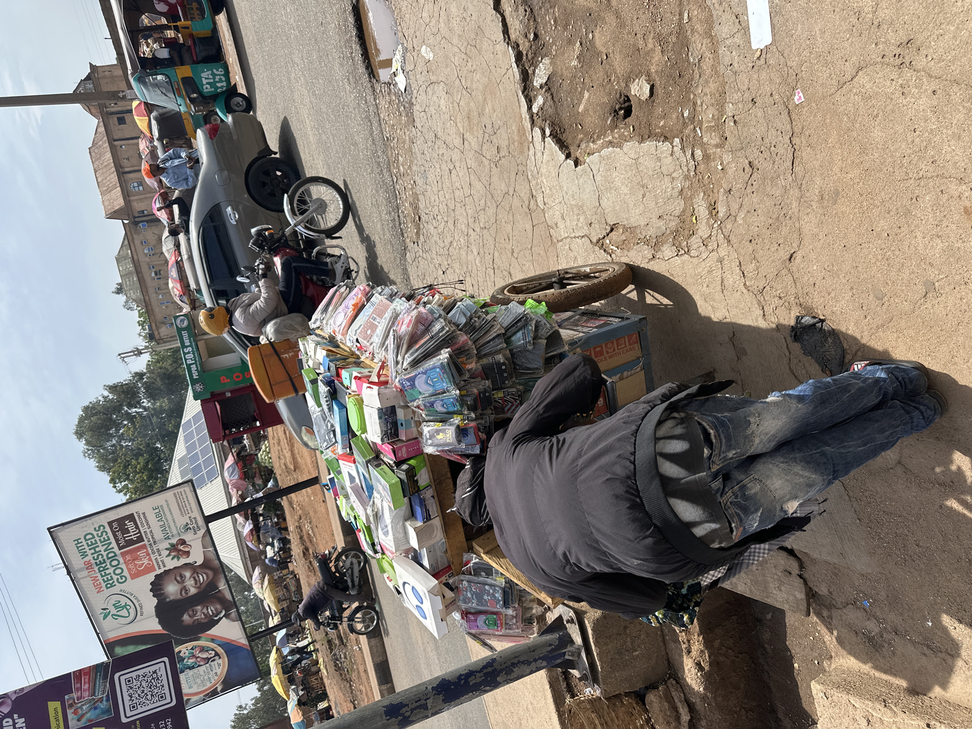 The image captures a vibrant street market scene in Gura Top, Nigeria, under bright daylight. In the foreground, a person wearing a black jacket and blue jeans is bent over, tending to a makeshift market stall. The stall is laden with numerous items, predominantly small electronics, phone accessories, and cases, many packaged in clear plastic or small boxes. A partially visible box on the stall has "DLE WITH CARE" printed on it.
Behind the vendor, the street is active with various vehicles, including a grey car, motorcycles, and a green and yellow auto-rickshaw (tricycle) bearing the text "PTA 2136". Other individuals are visible in the background, some on motorcycles and others near additional stalls, indicating a bustling commercial area.
The ground is characterized by cracked concrete and dusty, unpaved sections, with a small puddle reflecting the sky on the roadside. Strong shadows suggest it is a sunny day, likely mid-morning or afternoon.
A large billboard dominates the left side of the frame, advertising "Zyn REFRESHED GOODNESS REFINED SHEA BUTTER" for "Soft On Skin Moist On Hair" with various scent options like "PAPAYA | COCOA BUTTER | COCONUT | MINT | ROSE | TROPICAL SUNSHINE". It also states "AVAILABLE IN STORES NATIONWIDE" and lists contact information for "Illera Care Limited" (+2349099004400, www.zynshes.com). Below this, a smaller sign or billboard is partially visible with "PSPOA P.O.S OUTLET" and a QR code. Solar panels are noticeable on a building in the distant background. The overall scene depicts a dynamic urban or semi-urban marketplace.