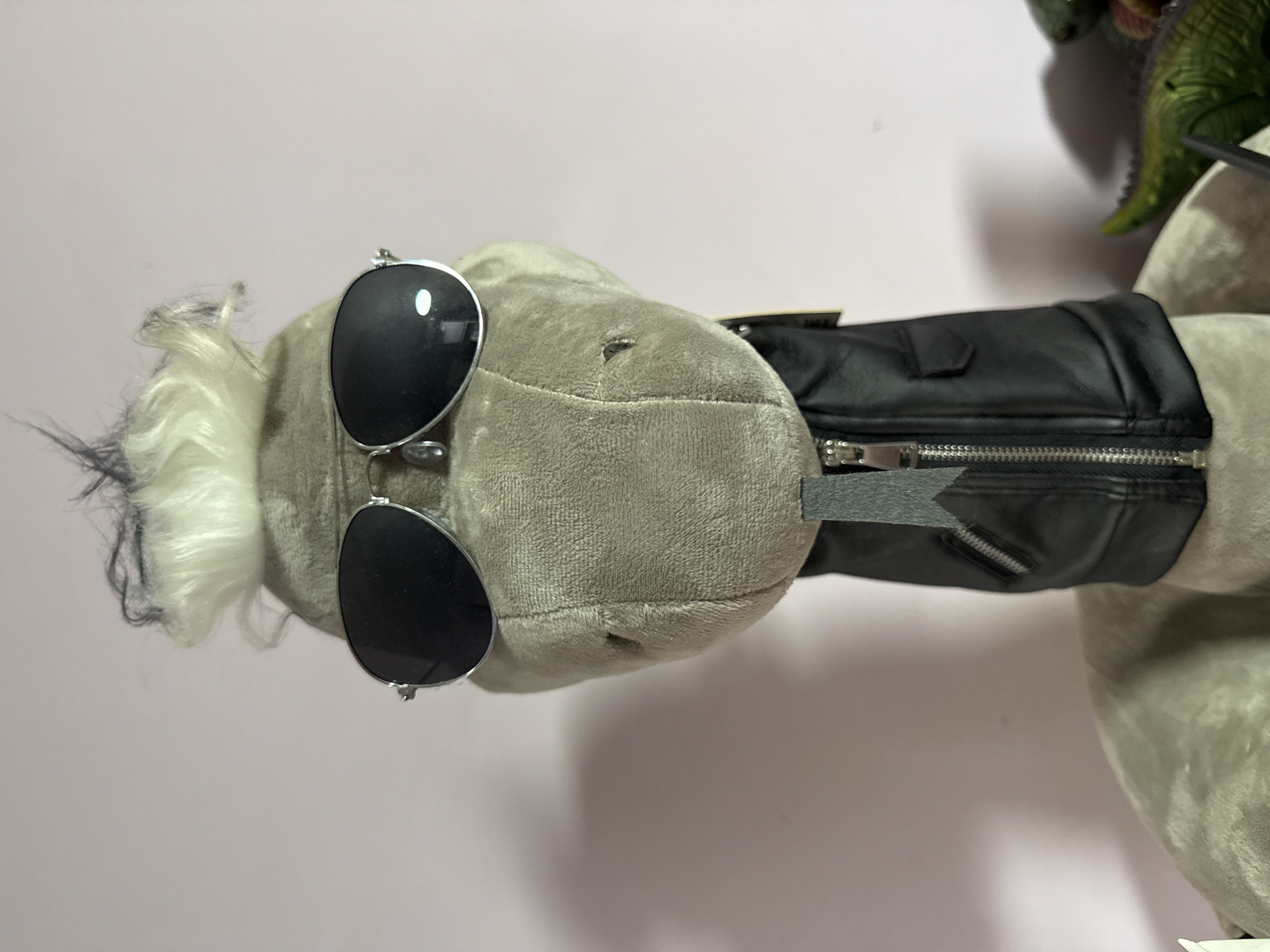 The image shows a close-up, vertically oriented view of a plush toy. The toy appears to be a gray creature with white and black hair styled around its head. It is wearing a black faux-leather jacket with a zipper and a gray fabric tag. Two dark, oval sunglasses are perched on the toy's head, one over each eye. The background is a plain, light pinkish-white surface, likely a wall or backdrop. There is no discernible activity or event taking place, and no specific time of day or weather can be determined. The setting is nondescript, offering no clear location cues beyond the possibility of being indoors. No text is clearly visible on the toy or in the background.