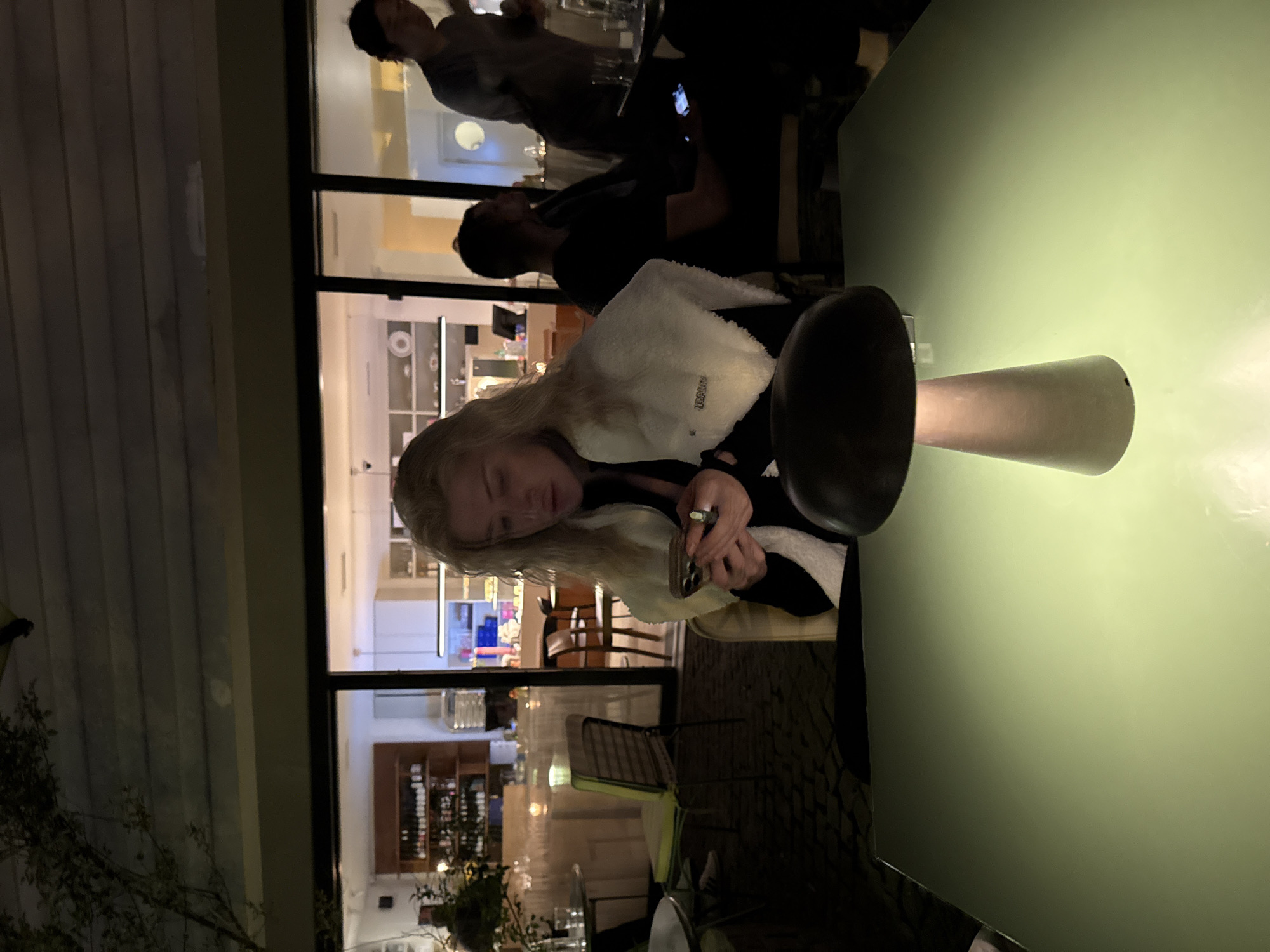 The image depicts an indoor/outdoor scene, likely at a restaurant or bar in Belgrade, Serbia, based on the presence of Cyrillic characters on interior signage (visible through the windows). A blonde-haired woman is seated at a table, looking at her cell phone. She is wearing a black shirt and a white fleece jacket. A mushroom-shaped table lamp is prominently visible on the table next to her. Through the glass windows behind her, other people are visible inside the establishment, one with a tablet or phone in his hand. A bar area stocked with bottles can also be seen. Outdoors, there are tables, chairs, plants, and lighting, suggesting a patio or terrace area. The time of day is likely evening or night, given the lighting conditions.