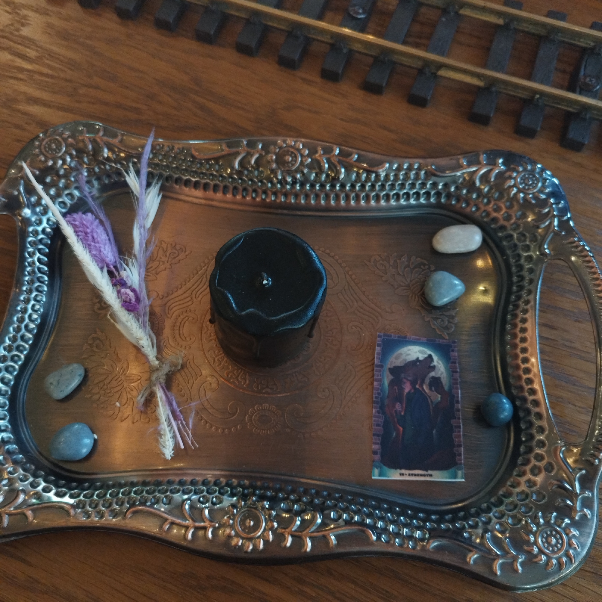 An overhead shot displays a still life arrangement on an ornate, metallic tray with a tarnished silver or bronze finish and intricate embossed patterns. The tray rests on a wooden surface, likely a table.
At the center of the tray is a small, black cylindrical candle, possibly unlit, with visible wax drips down its sides and a short wick. To the left of the candle, a small bundle of dried flowers, tied with twine, features white feathery stalks and small purple blossoms.
Scattered around the tray are five smooth, small pebbles of various shades, ranging from light grey to bluish-grey. To the right of the candle and flowers, a small card or print depicts an illustration under a full moon. The image shows two figures, one human-like and the other with distinct lupine features, against a background of brickwork. Visible text on the card reads "VI - STRENGTH".
In the upper background, out of focus, a section of model train tracks with dark ties and metallic rails is visible. The overall scene suggests a decorative or possibly symbolic arrangement, set indoors.