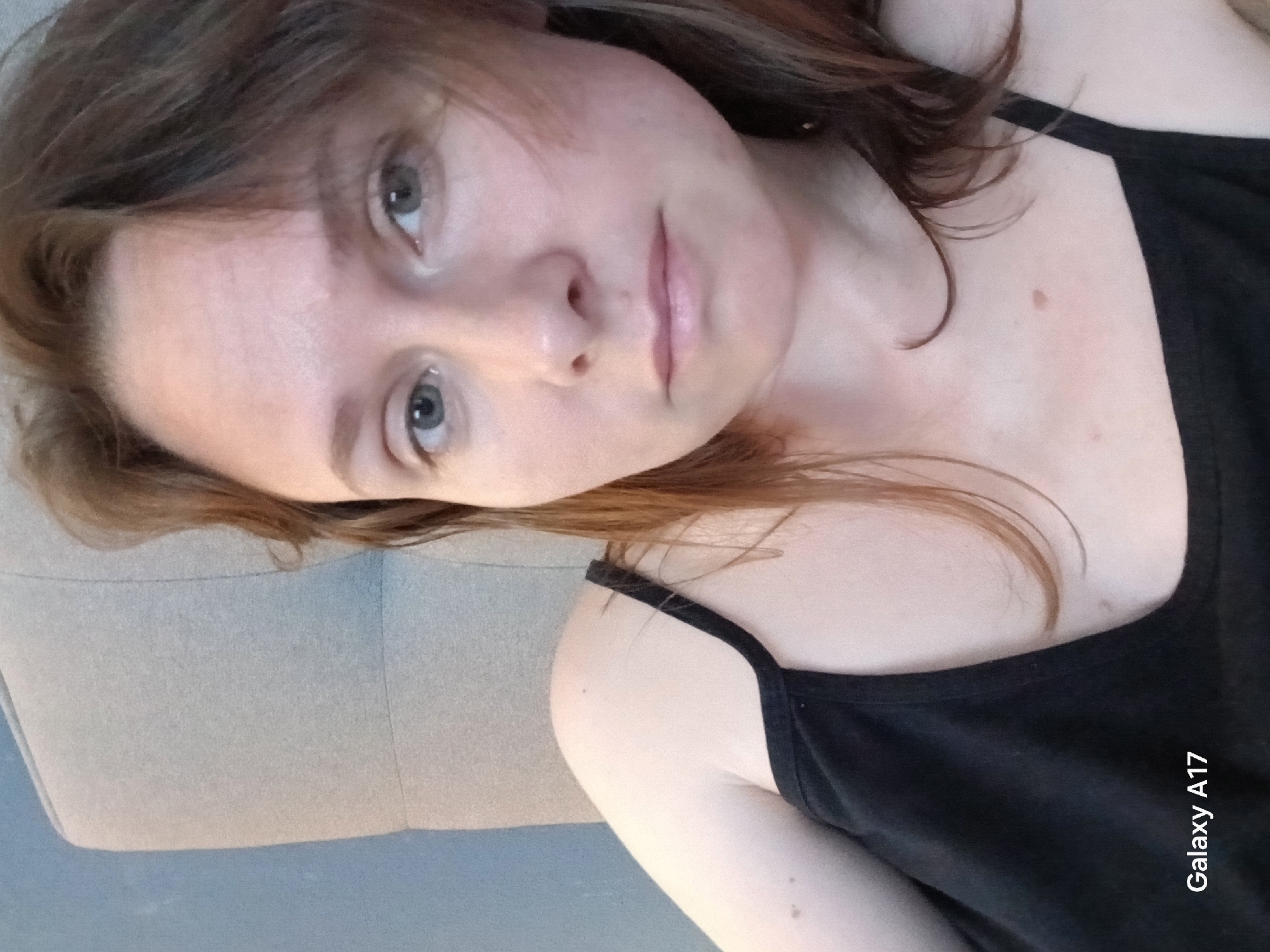 A young woman lies on a light gray surface. She wears a black tank top with thin straps. Her hair is brown with reddish highlights, partially obscuring her forehead. She has fair skin and light-colored eyes. A mole is visible on her shoulder. The image was taken with a Galaxy A17 phone.
