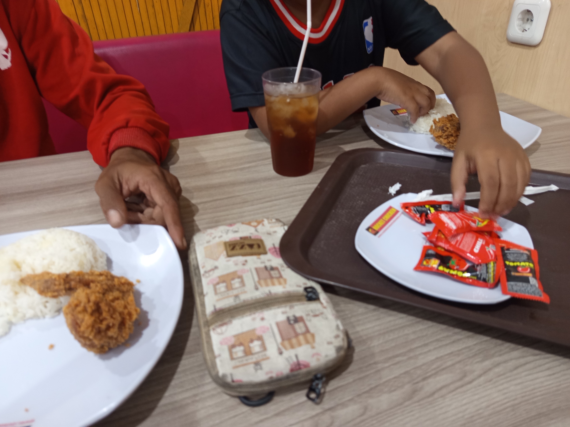 In Prajekan Lor, Indonesia, two individuals are seated at a wooden table, engaged in a meal. One person, wearing a red sweater, rests their hand on the table, while the other, dressed in a black t-shirt with a white trim and a logo, is reaching for food on a white plate. On the plate are servings of white rice and fried chicken. Adjacent to this plate is a brown tray holding another white plate laden with several small packets of red sauce. A clear plastic cup filled with iced tea and a straw stands between the two individuals. A small, patterned zippered pouch rests on the table near the first plate. The background features a pink booth seat and a wall with a yellow striped pattern, where a white electrical outlet is visible. The scene depicts a casual dining experience, possibly at a restaurant, with a focus on fried chicken and accompaniments.