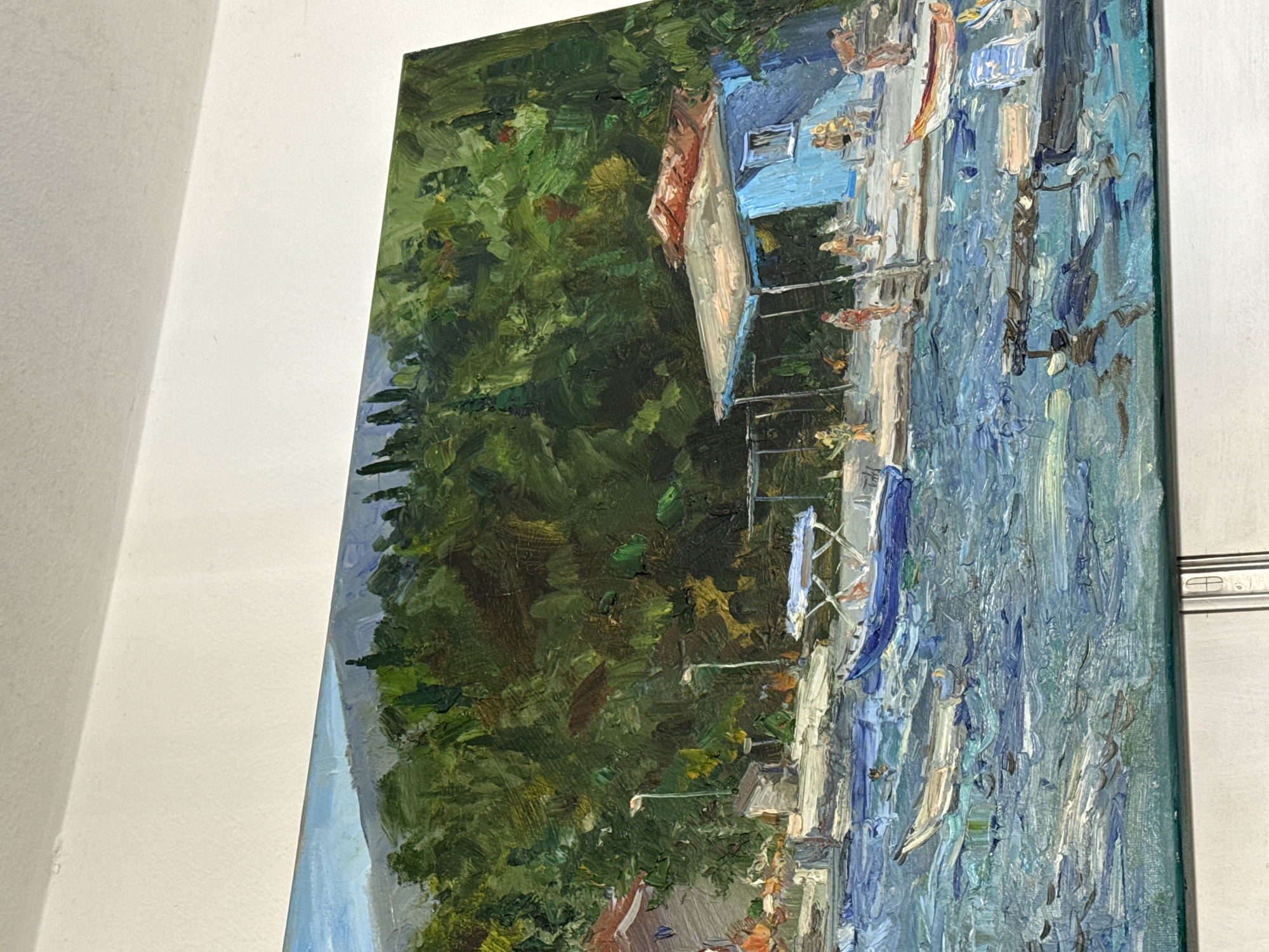 This is an oil painting depicting a waterfront scene. In the foreground, choppy blue water is rendered with thick impasto brushstrokes, suggesting movement and light reflection. Several small boats are visible on the water, including a long, narrow white boat and a larger blue boat with a canopy.
The middle ground features a paved promenade alongside the water. Along the promenade, there are several figures, some appearing to be walking or standing. A distinctive bright blue building with a reddish-brown roof is prominent on the right side of the promenade, featuring a window and an open doorway. Adjacent to it is a lighter-colored structure with a covered awning supported by poles. There are also streetlights and a small, shed-like structure.
The background is dominated by a dense, dark green forest that covers a hillside. In the far distance, hazy, muted blue mountains are visible beneath a pale blue sky. The painting appears to capture a sunny day with a lively atmosphere, likely a coastal town or village. The style is impressionistic, with visible brushwork and a focus on capturing light and color.
Given the visual elements of the painting, such as the blue sea, forested hills, and characteristic architecture of waterfront buildings, the scene strongly resembles locations along the Bay of Kotor in Montenegro. Herceg Novi, known for its coastal setting and proximity to forested hills, is a plausible location for such a depiction.