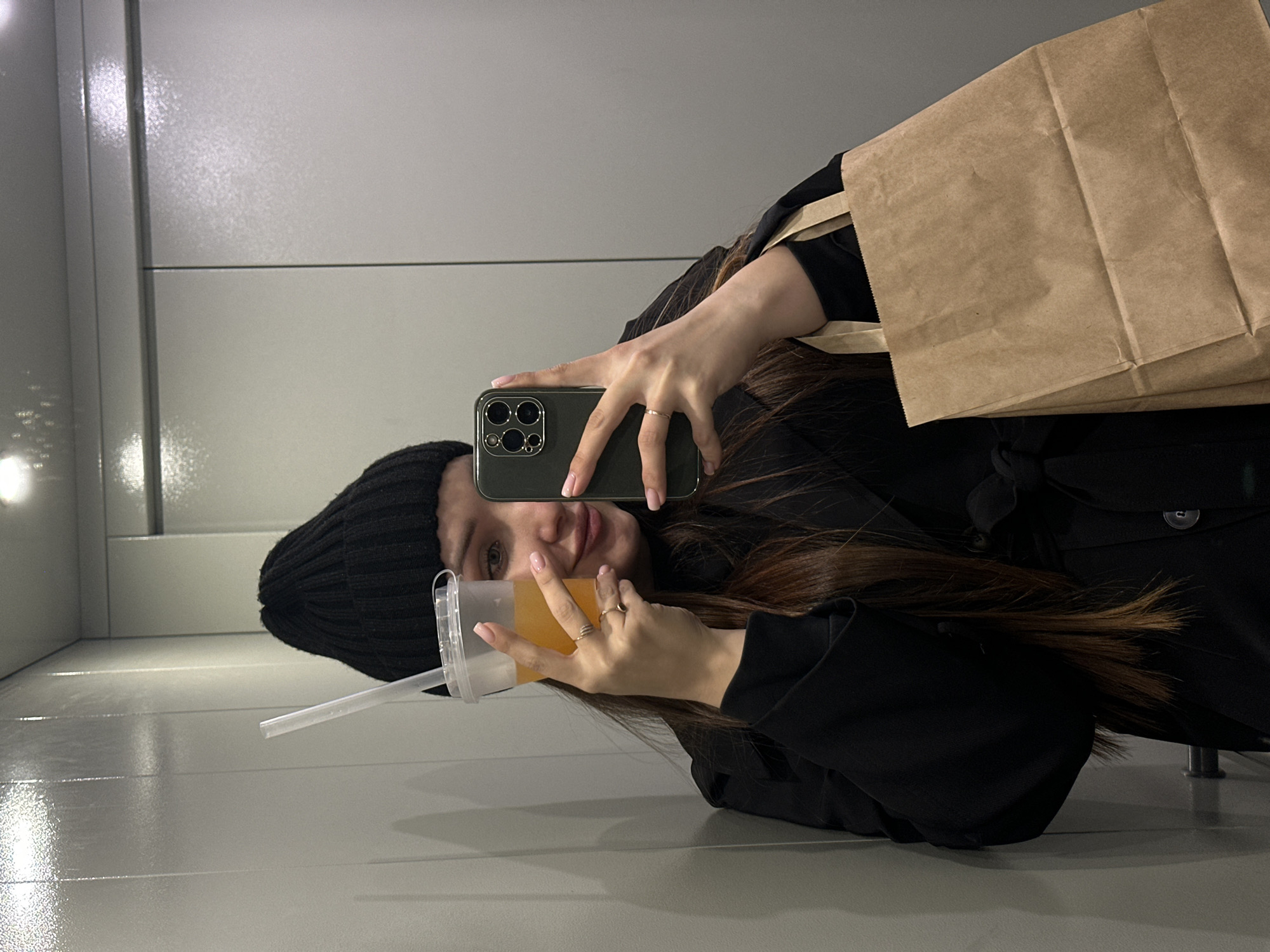 A young woman is depicted taking a selfie in what appears to be a reflective surface, such as a mirror or highly polished wall. She holds a dark-colored smartphone, featuring a triple camera system, in her right hand, positioned to capture her reflection. She is wearing a black beanie and a black long-sleeved top or coat. Her long, dark brown hair cascades over her shoulders.
She gazes directly at the camera with a gentle smile. In her left hand, she holds a clear plastic cup with a lid and straw, filled with a light orange-colored beverage. A crumpled brown paper bag with handles is slung over her left shoulder, partially visible behind her. She wears multiple thin rings on her left hand's ring finger and one on her right hand's ring finger. Her fingernails are painted with a light pink polish.
The background consists of plain, light grey or off-white panels, possibly reflective, with horizontal lines. The overall setting appears to be a minimalist indoor space with artificial lighting. The image is rotated 90 degrees clockwise. This scene is set in Chaikovsky, Russia. No discernible text is visible within the image.