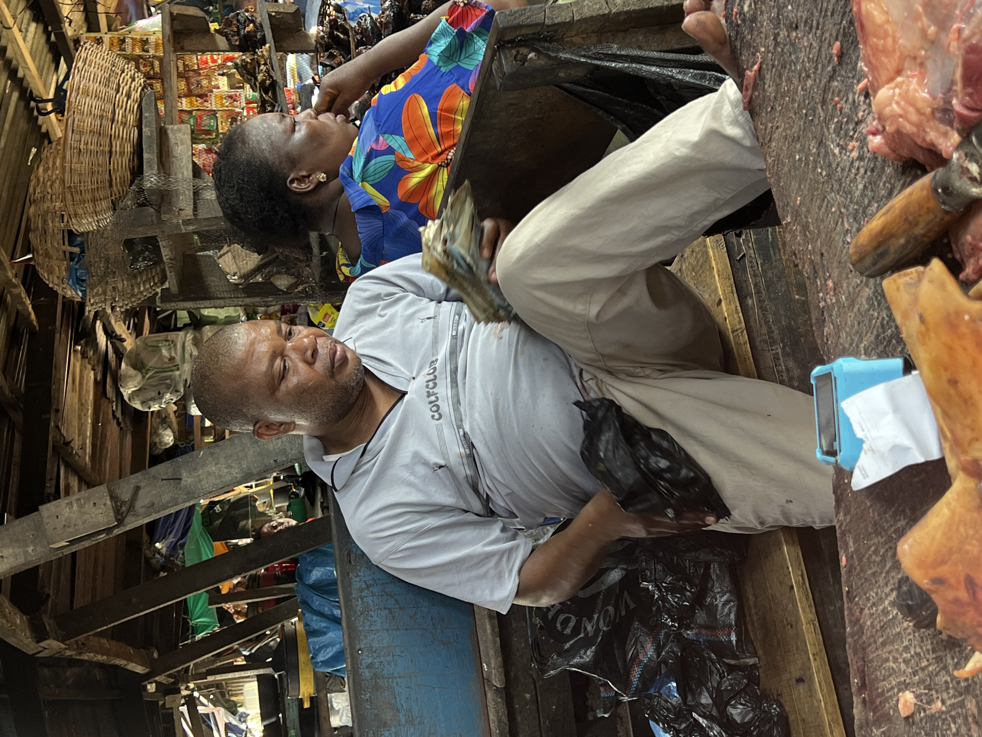 In an open-air market in Nkpor, Nigeria, a man in a light gray shirt with "GOLF CLUB" on the chest is seated on a blue-painted wooden structure with a black plastic bag underneath. He is holding a black bird. Nearby, a woman wearing a blue and floral top is seated. A wooden table is positioned close to them, on which there are pieces of meat, a knife, and a blue cash register. Woven baskets are visible in the background, and a wooden structure with various items is above the individuals.