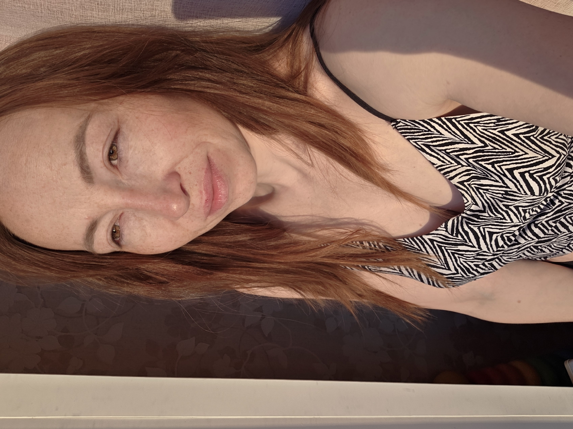 The image, rotated 90 degrees clockwise, displays a woman with long, reddish-brown hair, relaxing indoors in Chaikovsky, Russia. She is depicted from a slightly elevated viewpoint, looking towards the camera with a gentle, contented smile. Her fair skin features visible freckles. She is wearing a sleeveless top with a prominent black and white zebra pattern and thin black straps. Strong natural light, likely sunlight, illuminates her face and hair, casting distinct shadows on her upper arm and the surrounding areas, suggesting a daytime setting, possibly morning or afternoon. The upper background shows a light-colored, textured fabric, resembling a pillow or blanket. Below her, a dark brown surface with a subtle floral or leafy pattern, likely wallpaper, is visible, while a plain white surface forms the very bottom of the frame. The overall scene conveys a sense of calm. No text is visible in the image.