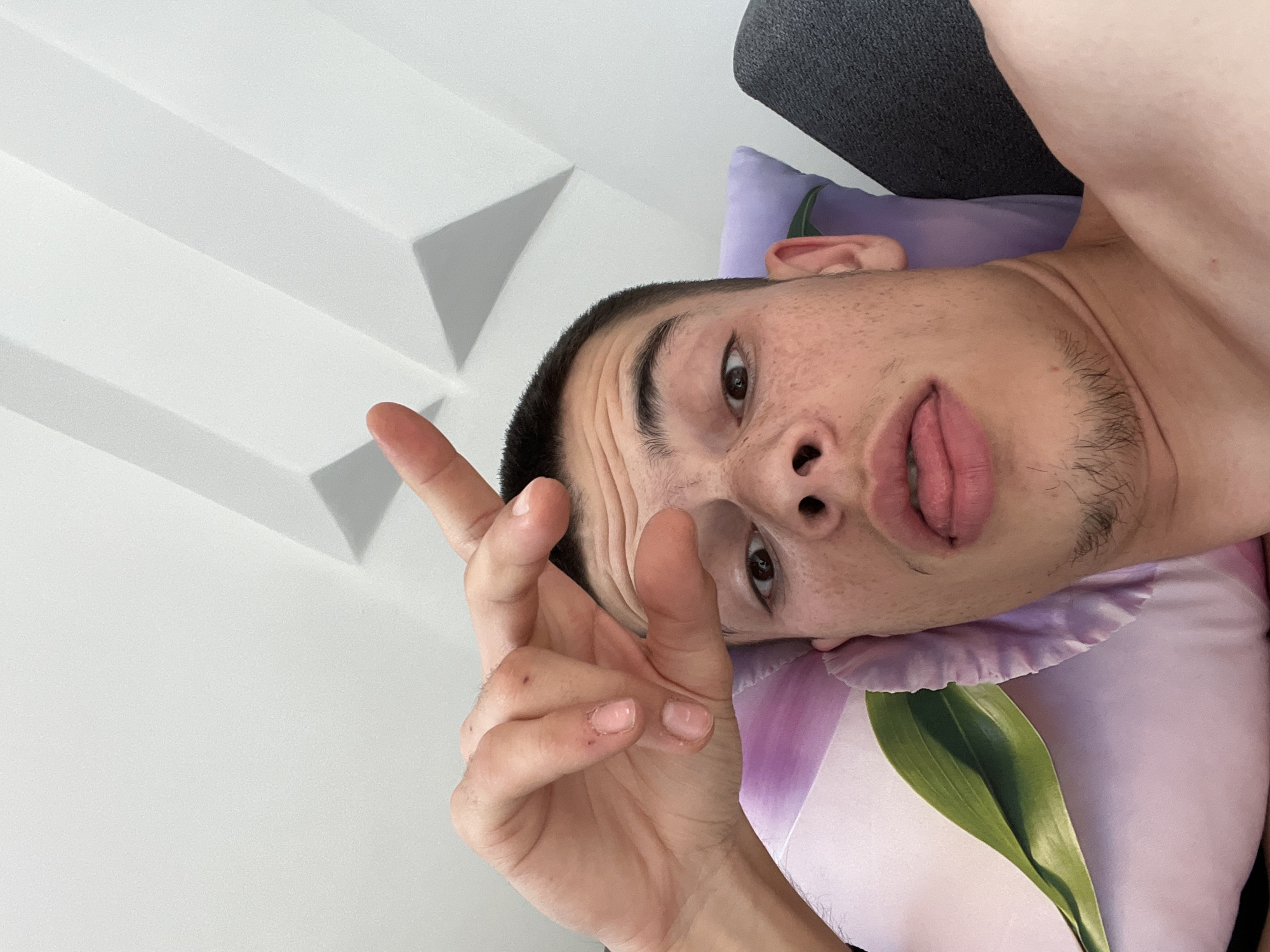 A close-up, selfie-style shot shows a young man lying down, with his head and upper torso filling the frame. He is looking towards the camera with his eyes wide open. His right hand is raised, with his index finger and thumb forming a small circle, held near his forehead. His tongue is sticking out slightly. He has short dark hair and a light stubble on his chin and upper lip. He has a smattering of freckles across his nose and cheeks. He is lying on a pillow with a floral design, and a dark gray object, possibly a sofa, is visible to his right. The background consists of a white, angled wall with geometric patterns and shadows. This appears to be an indoor setting. There is no information in the image that indicates the location is the city of Karavelovo, Bulgaria.