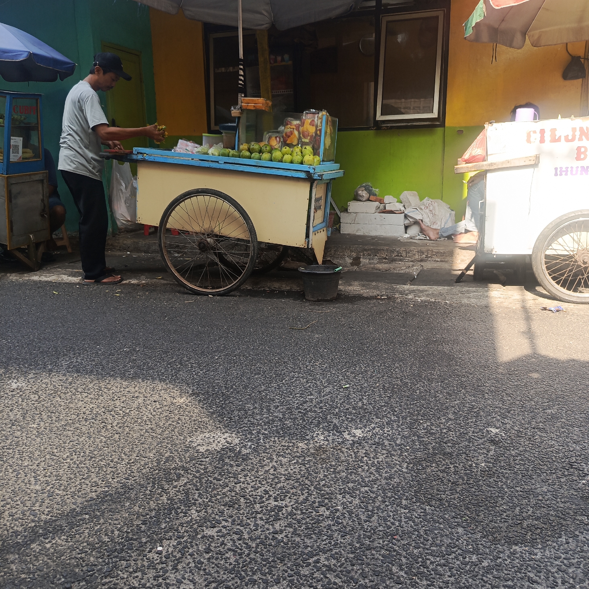 A man wearing a light grey t-shirt, black pants, and a black baseball cap stands next to a fruit cart on a street in South Jakarta, Indonesia. He is holding some green fruit in his hands. The cart is light yellow with a blue trim and has a large spoked wheel. In front of the cart, there are several green fruits displayed. Behind the cart, there is a colorful wall, painted green on the lower half and yellow on the upper half. To the right of the fruit cart, another vendor cart is partially visible, painted white with some text and a pink umbrella. In the background, a building with windows is visible. The ground is paved with asphalt.