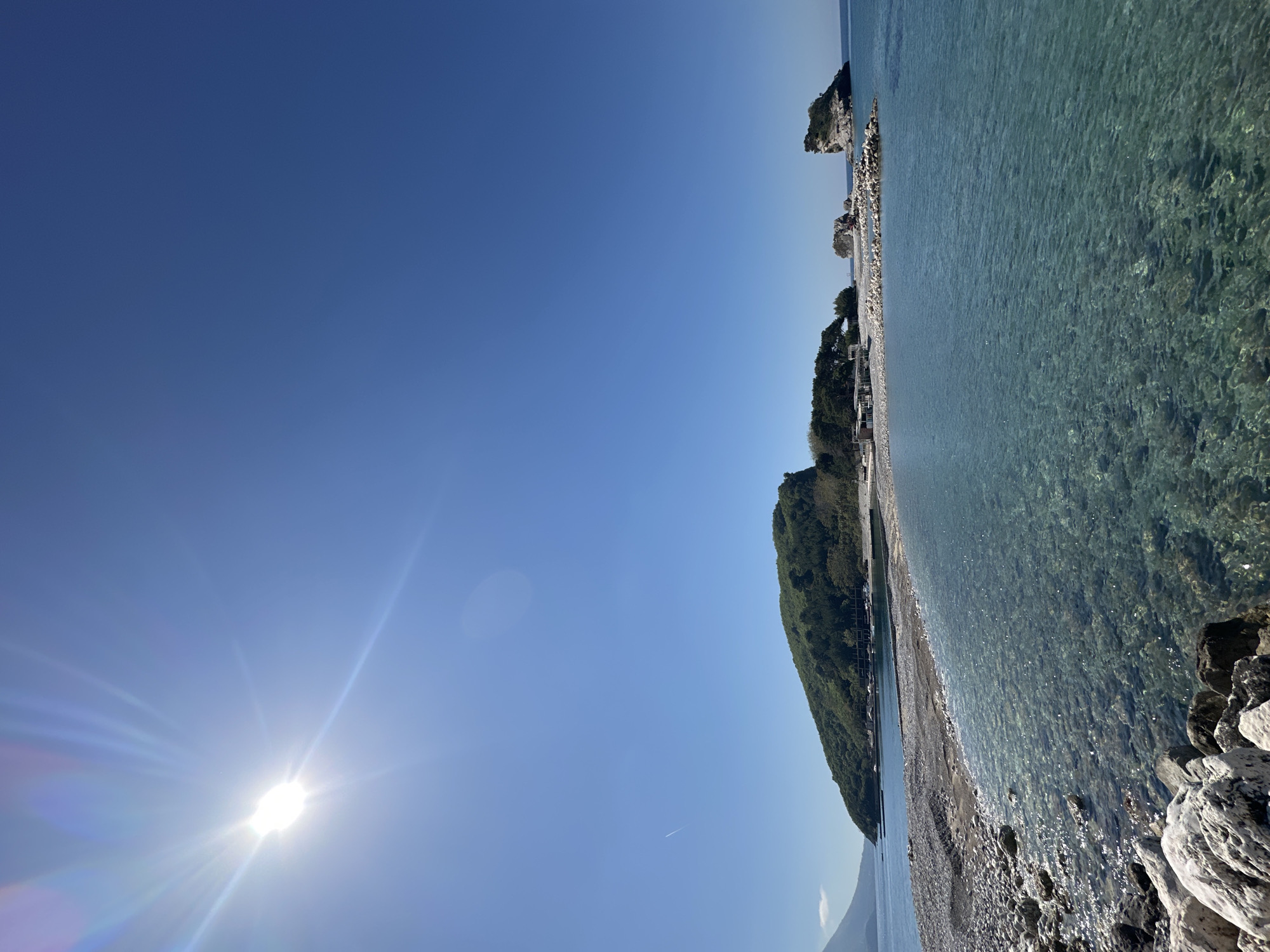 The image shows a shoreline in Budva, Montenegro, during daylight. The sky is clear and blue with the sun visible at the bottom left corner of the frame. The shoreline is to the right with clear water and rocky coastline. Buildings and trees are visible inland.