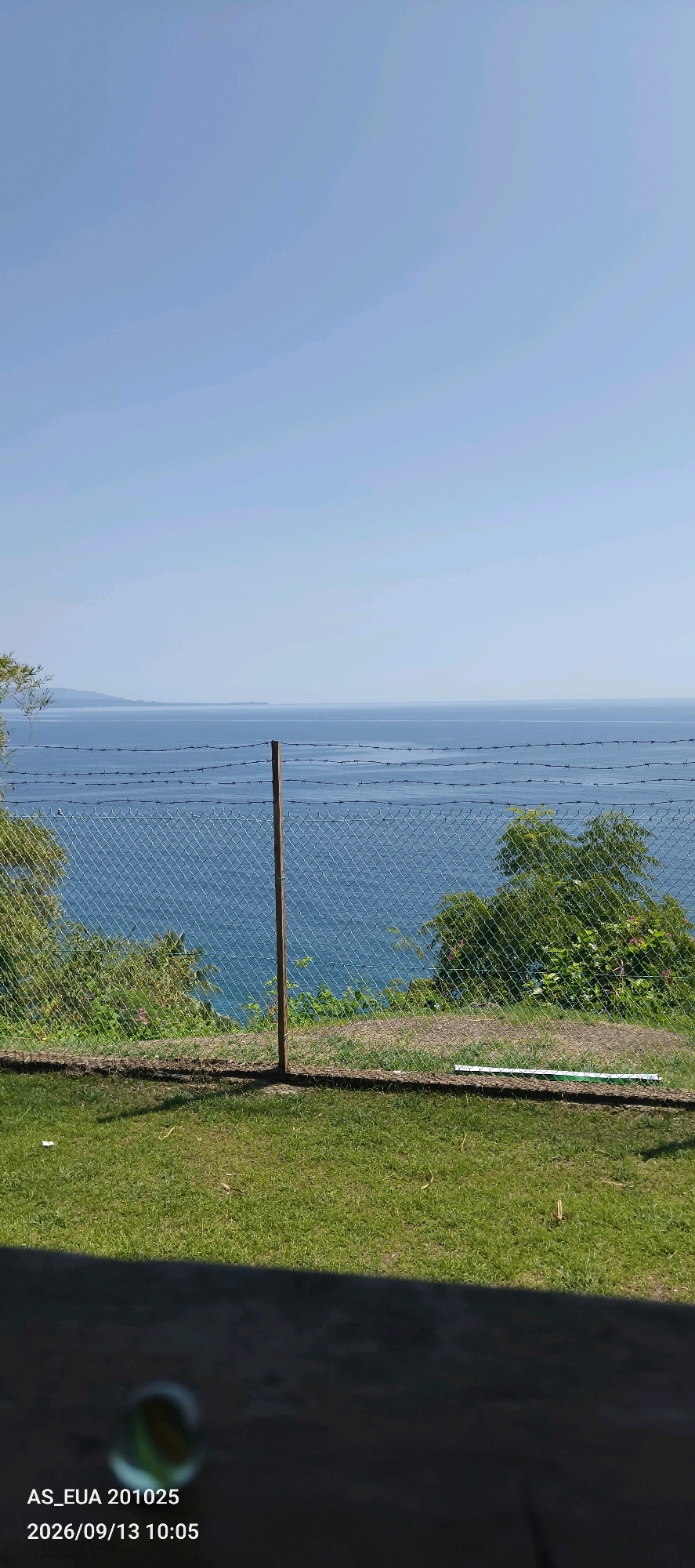 A wide, horizontal shot captures a serene coastal view from a grassy embankment in Wewak, Papua New Guinea. The bright, clear sky transitions into the calm, blue expanse of the ocean, meeting a distant, faint shoreline on the horizon. A chain-link fence with a single wooden post bisects the scene horizontally, with strands of barbed wire strung above it, suggesting a boundary. Lush green vegetation covers the embankment, with trees visible on either side of the fence. The foreground features a well-maintained lawn, with a shadowy area in the bottom left corner revealing a dark, textured surface, possibly a table or ledge, and a partially visible glass marble. Text overlays in the bottom left corner indicate "AS_ EUA 201025" and "2026/09/13 10:05".