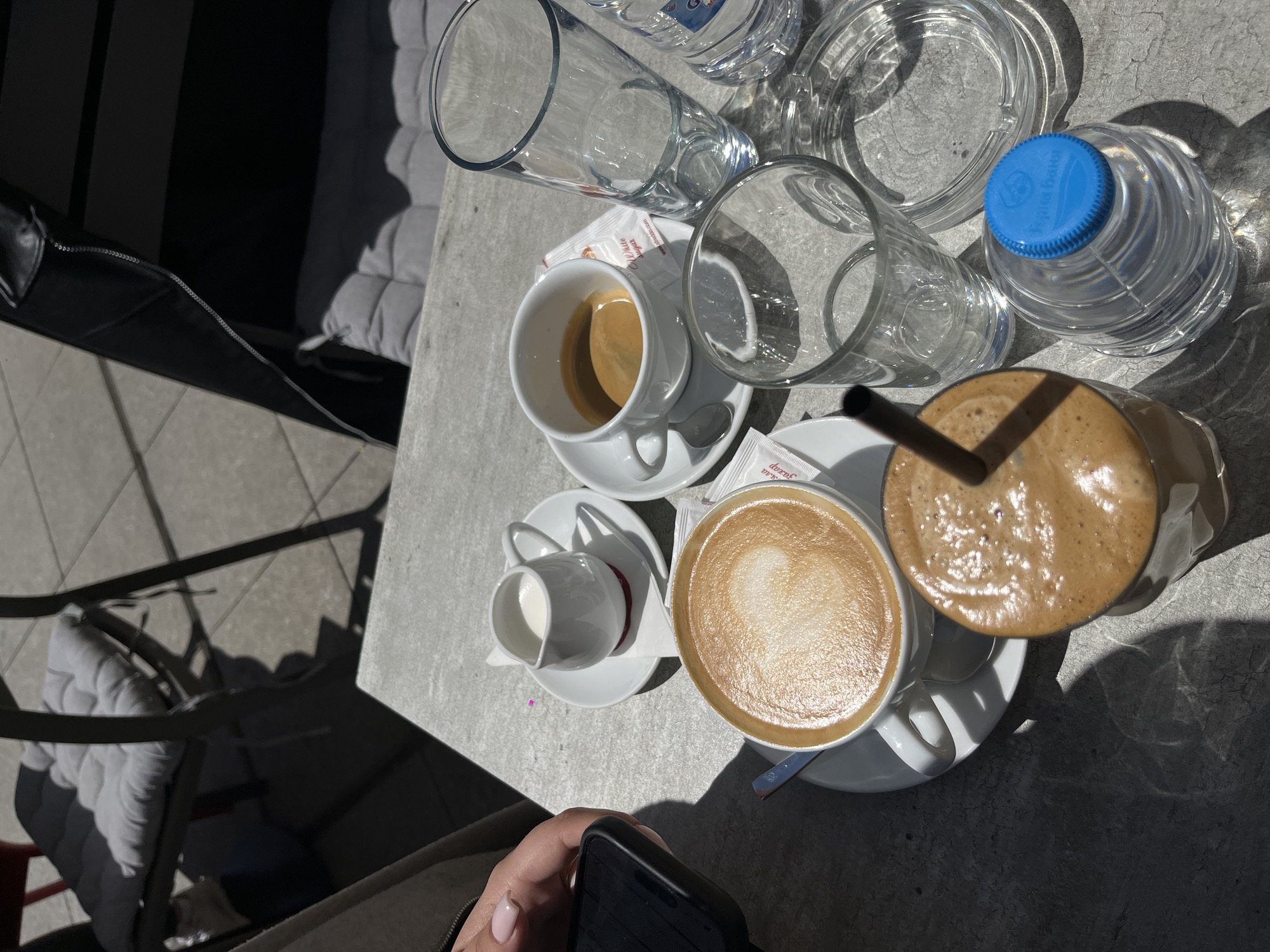 The image depicts a table setting on what appears to be a sunny day. The location is likely outdoors, possibly a cafe or restaurant in the city of Plovdiv, Bulgaria, based on the bottle of вода with Cyrillic lettering and the general aesthetic.
On the table, there are four cups of coffee, each with a saucer: an espresso, a cappuccino, a coffee with milk (or a cup of milk), and a blended coffee beverage in a tall glass with a straw. Adjacent to the coffees are several empty water glasses and a sealed bottle of water. Packets of sugar are also visible.
In the lower part of the image, a hand is partially visible holding a black smartphone.
The table is made of a grey concrete-like material. There is a chair with a patterned cushion to the left of the table.