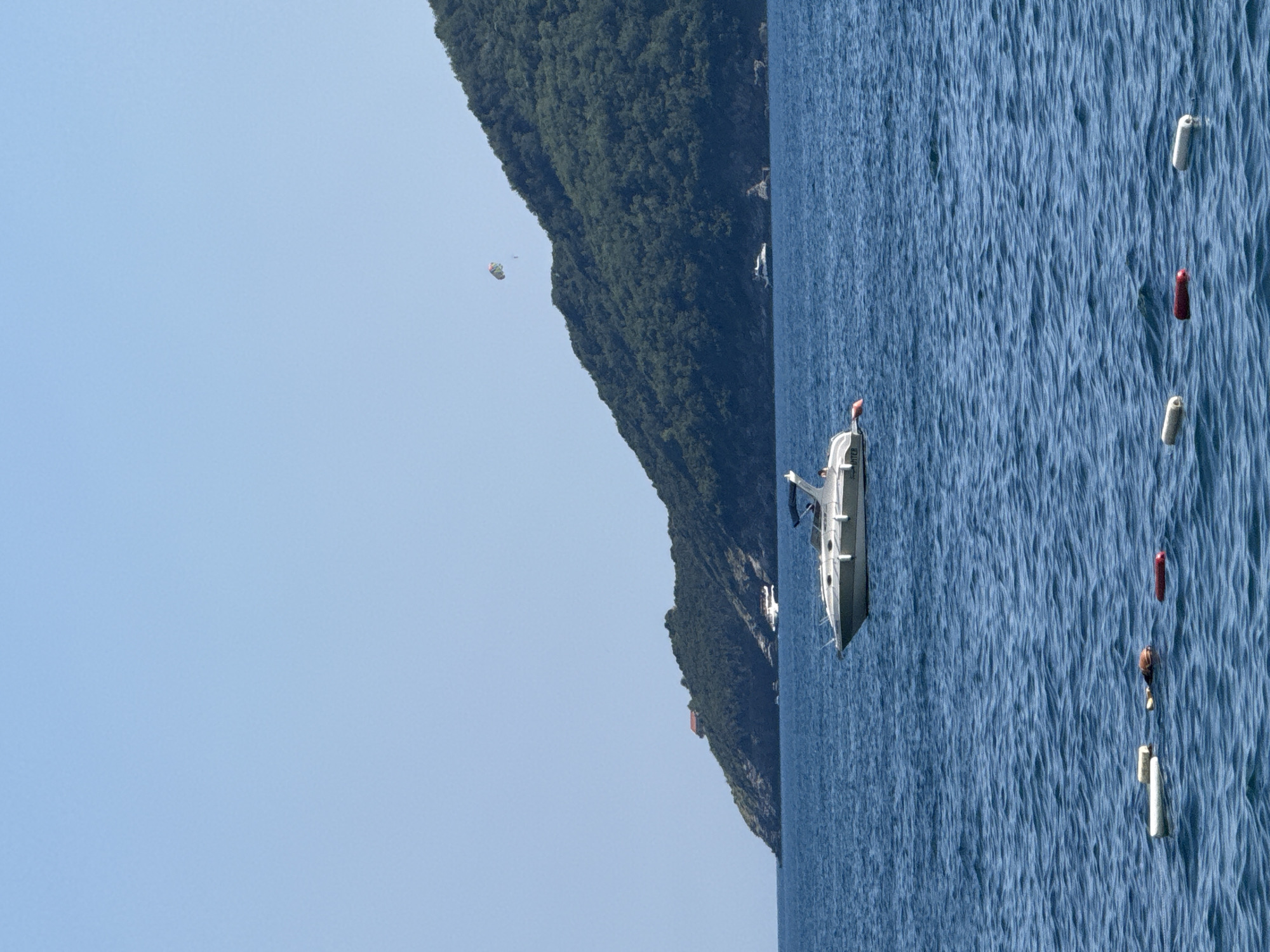 The image depicts a scene on the water near the coast of Budva, Montenegro. In the foreground, several white and red buoys are floating on the blue, rippling water. A white motorboat is positioned in the middle ground, with its bow facing left and a dark blue canopy over the seating area. In the background, a lush green, tree-covered hillside slopes down towards the water. A parasail with a person attached is visible in the sky above the hillside. A smaller white boat is anchored in the distance near the shore, and a reddish-brown structure is visible on the crest of the hill. The sky is a clear, light blue.