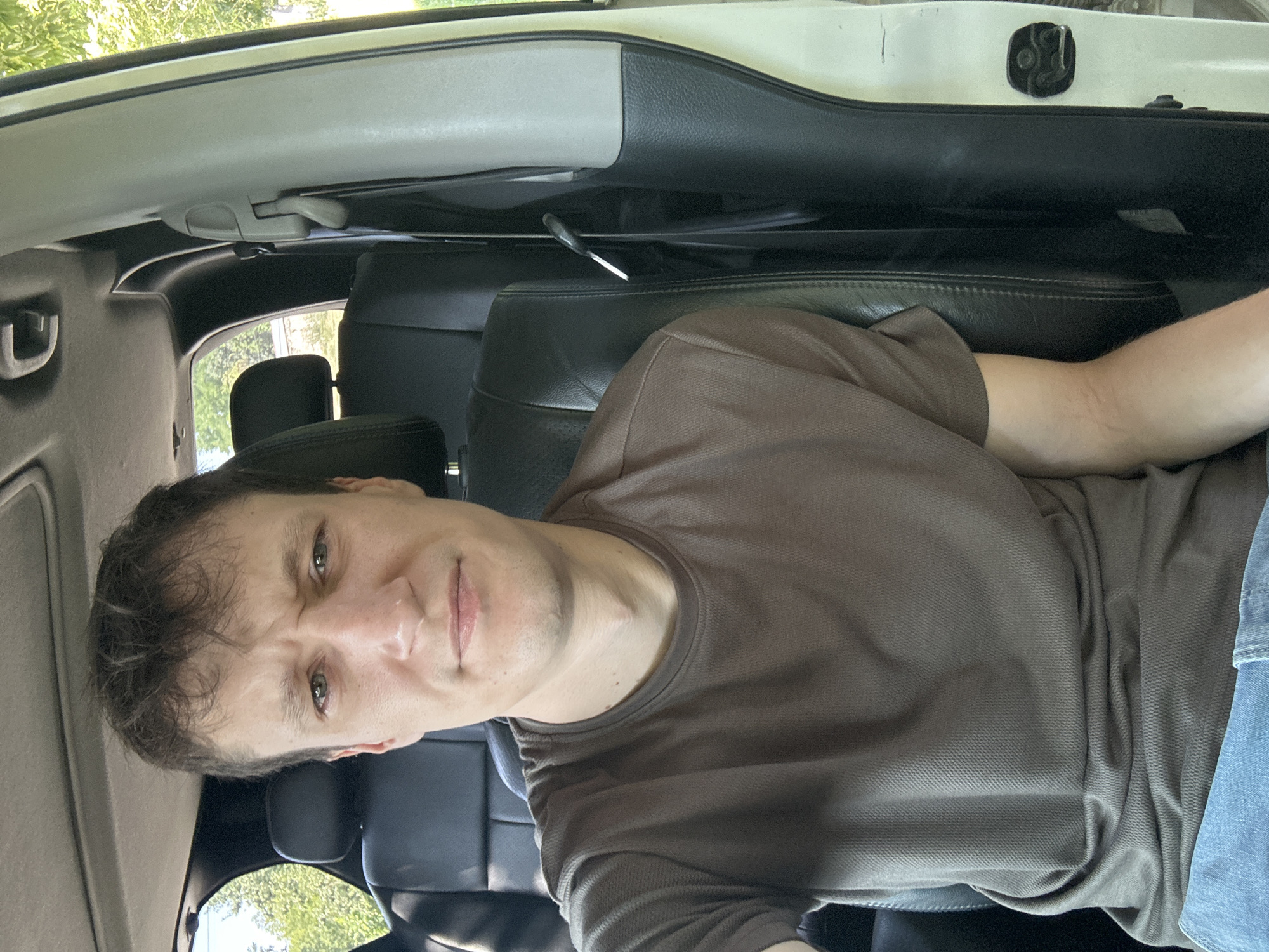 The image shows a man sitting in the driver's seat of a vehicle, possibly a car or an SUV, with the door open. He is looking towards the camera with a furrowed brow and his mouth slightly open. He is wearing a brown t-shirt and blue jeans. The interior of the vehicle appears to be black leather seats. Sunlight is filtering in from outside, suggesting it is daytime. The background shows trees and foliage, indicating an outdoor setting, potentially in a rural or suburban area, but not specifically Makiv, Ukraine.