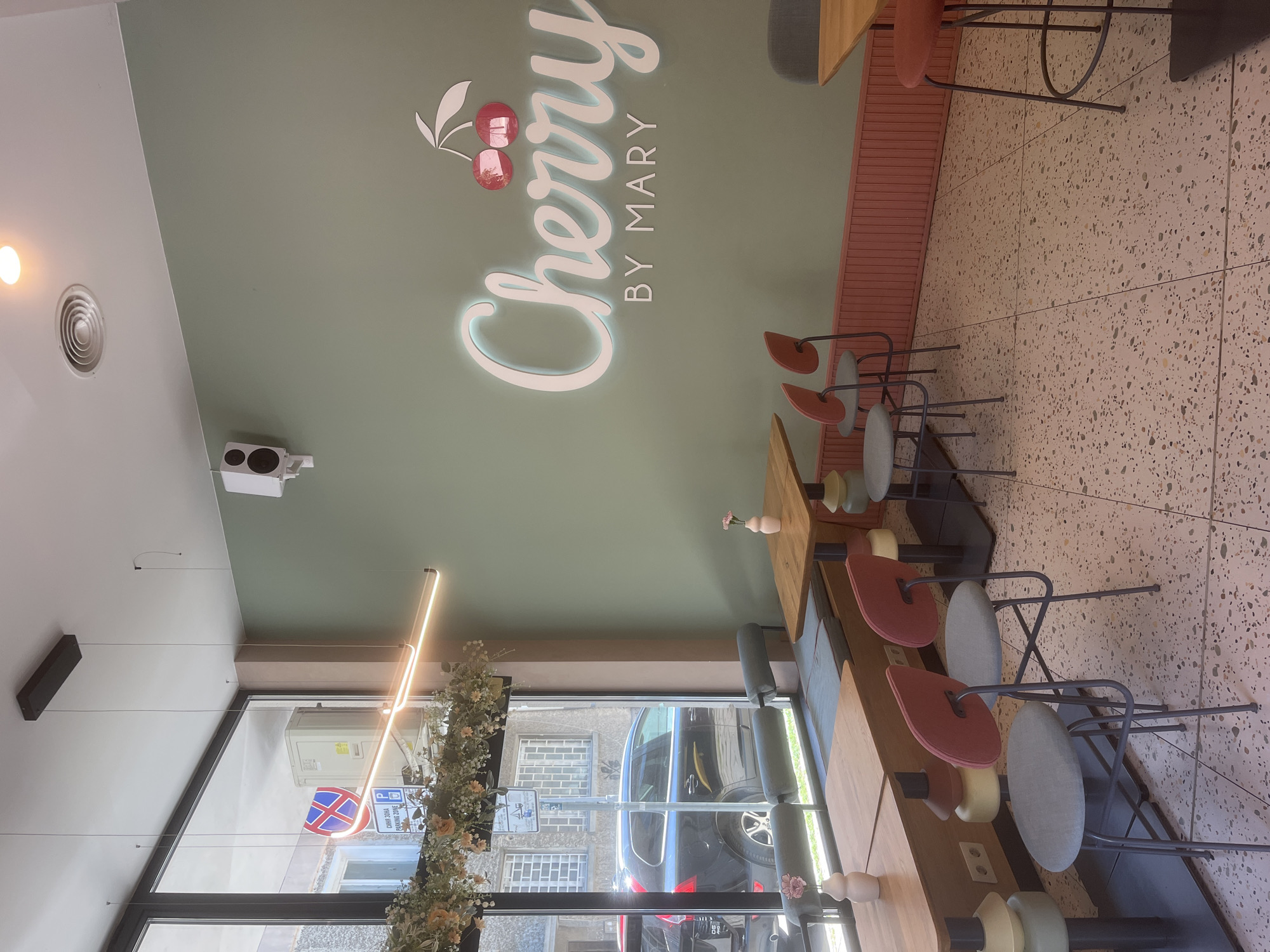 The image shows the interior of a cafe or restaurant named "Cherry by Mary" in Varna, Bulgaria. The interior features a light green wall with the cafe's name in stylized lettering. There are tables and chairs arranged along the right side of the image, with a window running along the bottom. Through the window, a street scene is visible, including a car and buildings. The lighting is a mix of natural light from the window and artificial lighting from fixtures on the ceiling. The floor is a patterned, terrazzo-style tile. There are decorative elements such as hanging foliage and a sign with parking symbols visible through the window.