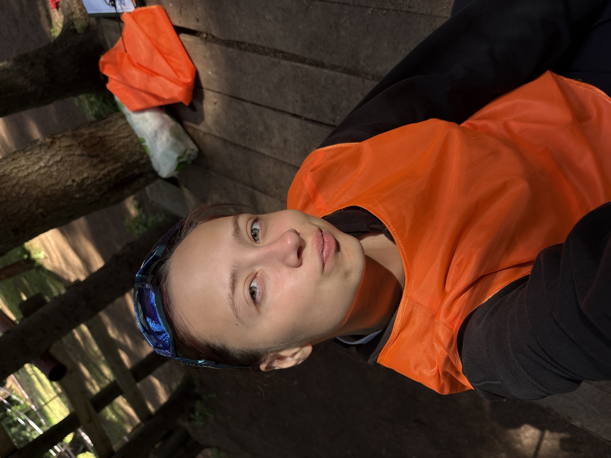 The image shows a young person wearing an orange vest and blue-tinted sunglasses, looking up towards the camera. Their expression is neutral, with slightly pursed lips. The person is outdoors, on a wooden surface that appears to be a deck or boardwalk. In the background, there are trees and sunlight filtering through the branches, suggesting it is daytime. To the left, there are some orange objects that look like folded bags or tarps, and a white and green object that could be a rolled-up mat or sleeping bag. The lighting suggests it might be a sunny day, possibly in the late afternoon given the shadows. The setting appears to be a natural or park-like environment. There is no visible text in the image.