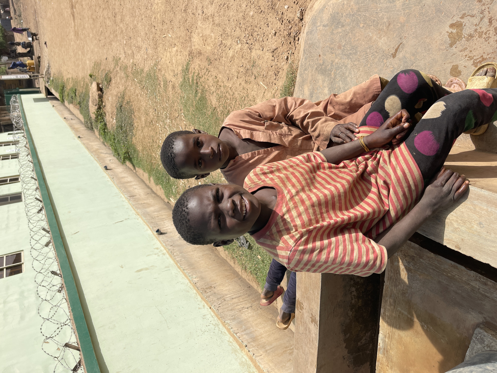 Two children are lying on a concrete ledge outdoors. One child is wearing a red and tan striped shirt and black leggings with large colorful polka dots. The other child is wearing a solid tan long-sleeved shirt and similar polka-dotted leggings. Both children are dark-skinned. One child is smiling broadly. The other looks serious. There is a building with barbed wire on top of it in the background. Beyond the ledge is a dirt area with sparse greenery. The location is Chikaji, Nigeria.