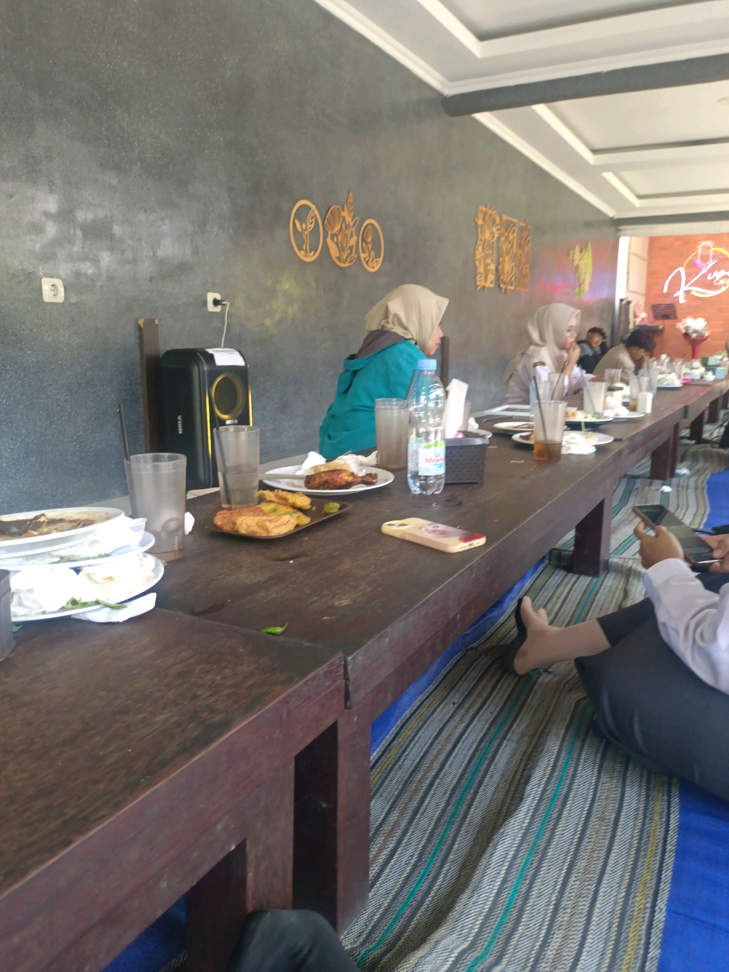 In Kerangkulon, Indonesia, a group of people are dining at a long, wooden table in what appears to be an outdoor or semi-outdoor cafe. The table is laden with plates of food, some partially eaten, as well as drinks in glasses and bottles. The individuals, some wearing hijabs, are seated on the floor on a blue mat with a striped pattern, suggesting a casual dining atmosphere. The background shows a textured gray wall with decorative wooden cutouts and a neon sign that is partially visible. The lighting suggests it is daytime.