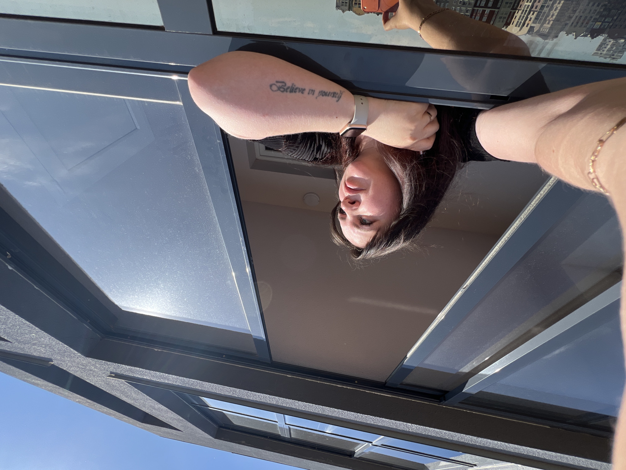 A woman with dark hair and fair skin is looking into the camera, with a portion of her body appearing in the frame. She has a tattoo on her left forearm that reads "Believe in yourself" and is wearing a wristwatch on the same wrist. A ring is visible on a finger on her right hand. The reflection of city buildings is visible on a window above her. The sky is blue. The location appears to be indoors, looking upwards through a glass structure. No specific information indicates the city of Pervomaysky, Russia.