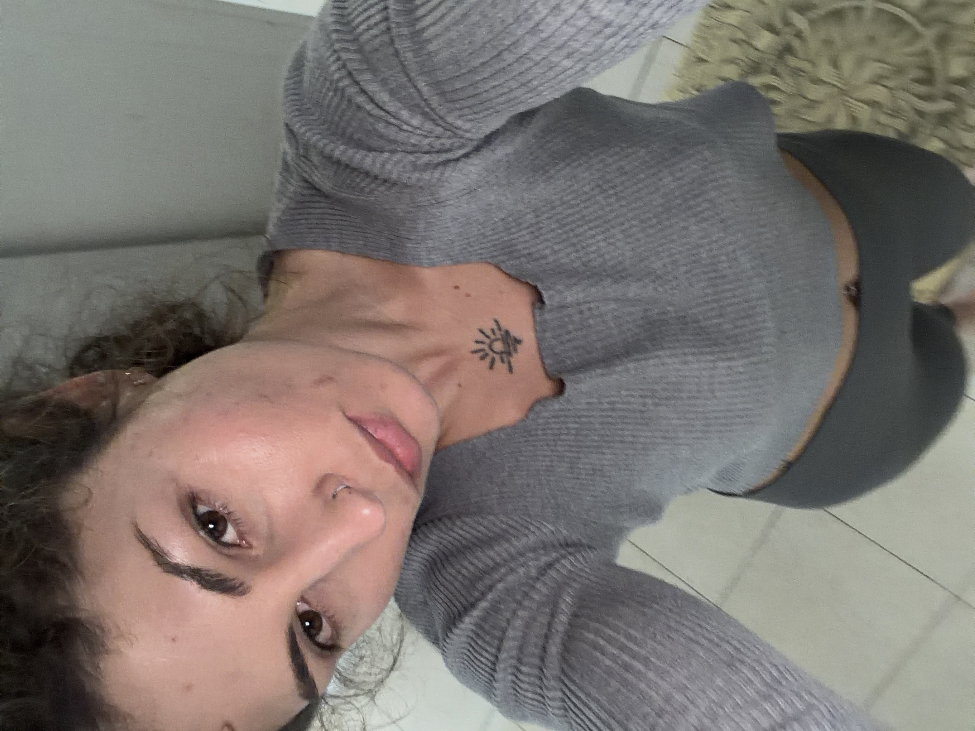 A medium shot of a woman with curly hair, brown eyes, and a nose piercing, wearing a gray long-sleeved, rib-knit crop top and gray leggings. She has a sun-shaped tattoo on her upper chest. The background shows a carpet and tiled flooring. The location and specific actions are not discernible from the image. The image does not provide the location in Kiryat Bialik, Israel.