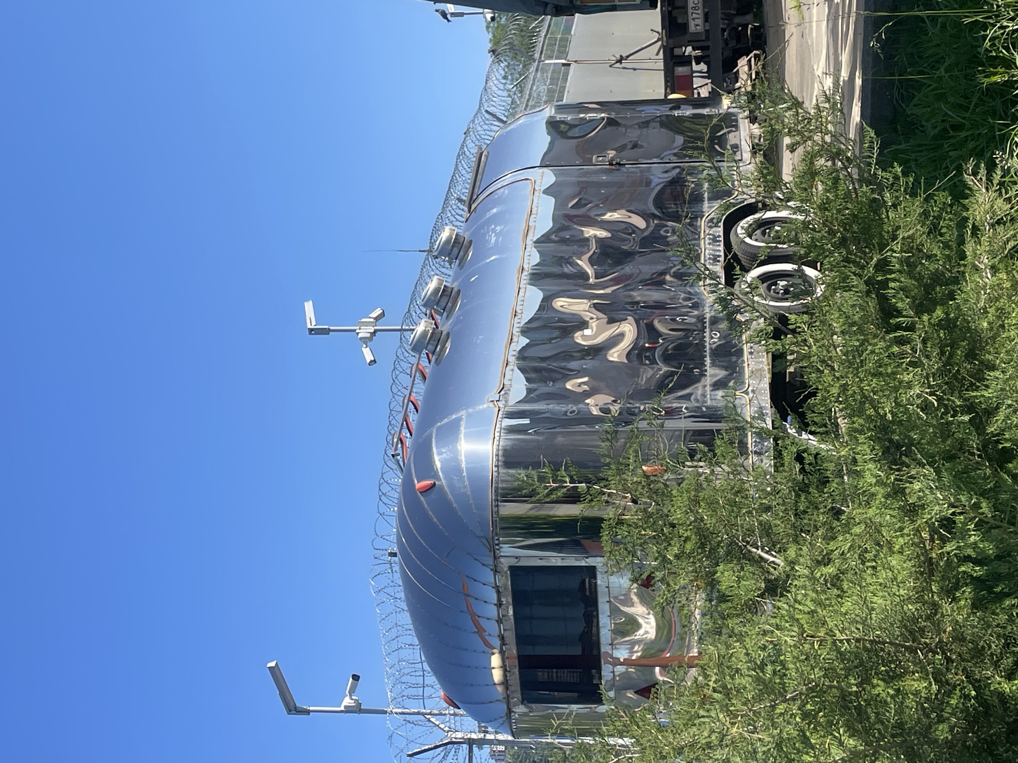 The image, rotated 90 degrees counter-clockwise, displays a highly polished, metallic, custom-built travel trailer, possibly an Airstream, situated outdoors under a clear, bright blue sky in Sochi, Russia. The trailer's reflective surface prominently mirrors its surroundings, showing distorted wavy patterns of the blue sky, green foliage, and other shapes.
A key feature of the trailer is its robust security system. Concertina wire (razor wire) is strung along the top perimeter, and multiple security cameras are mounted on poles around and possibly on the trailer itself. One visible pole on the left supports two cameras, while additional cameras and antennas are seen on the roofline. Dense green vegetation, including bushes and small trees, partially covers the lower right side of the trailer, obscuring its wheels, which appear to have white sidewall tires.
In the upper right background, part of a larger vehicle, likely a truck, is visible, showing a partial license plate with "y 178c" discernible. The scene is illuminated by bright daylight, indicating sunny and clear weather. No human activity is present, highlighting the secured and static nature of the setup. The overall impression is one of a uniquely customized and heavily protected mobile unit.