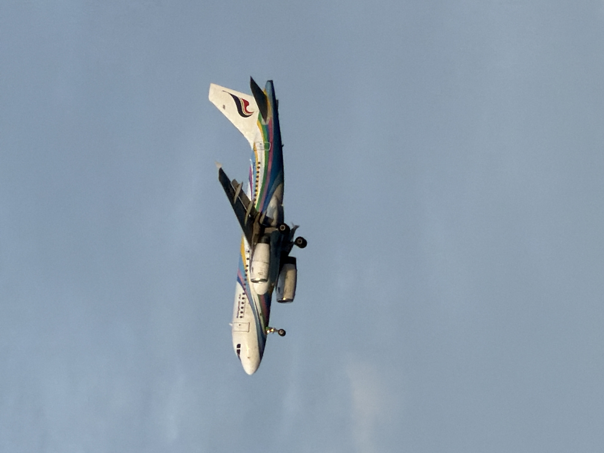 An airplane, with the tail logo of Bangkok Airways, is captured in mid-air against a pale blue sky with faint clouds. The aircraft's landing gear is extended, indicating it is likely in the process of landing or taking off. The plane is white with a colorful striped pattern along its fuselage and features two engines mounted under its wings. The scene is set with the plane positioned towards the upper right quadrant of the frame. The provided contextual information places this image in Ko Samui District, Thailand, suggesting the aircraft is operating within or around the vicinity of the island's airport.