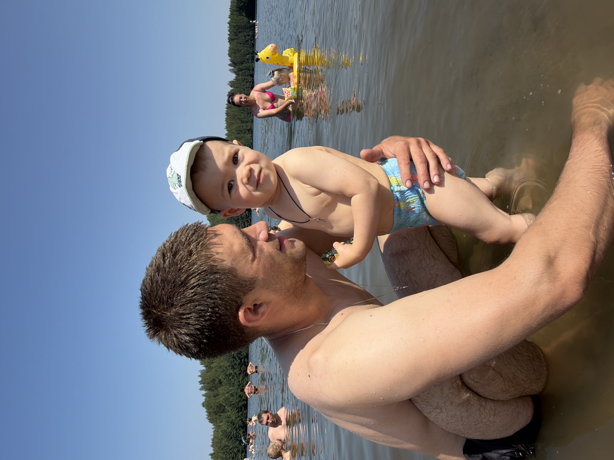 In Chernushka, Russia, a man and a young child are in a body of water. The man, who appears to be the father, is holding the child. The child is wearing a diaper and a cap. In the background, other people are visible in the water, some swimming and others using inflatable toys. The water is calm, and the sky is clear and blue. Trees line the shore in the distance. The scene depicts a recreational activity on a warm, sunny day.
