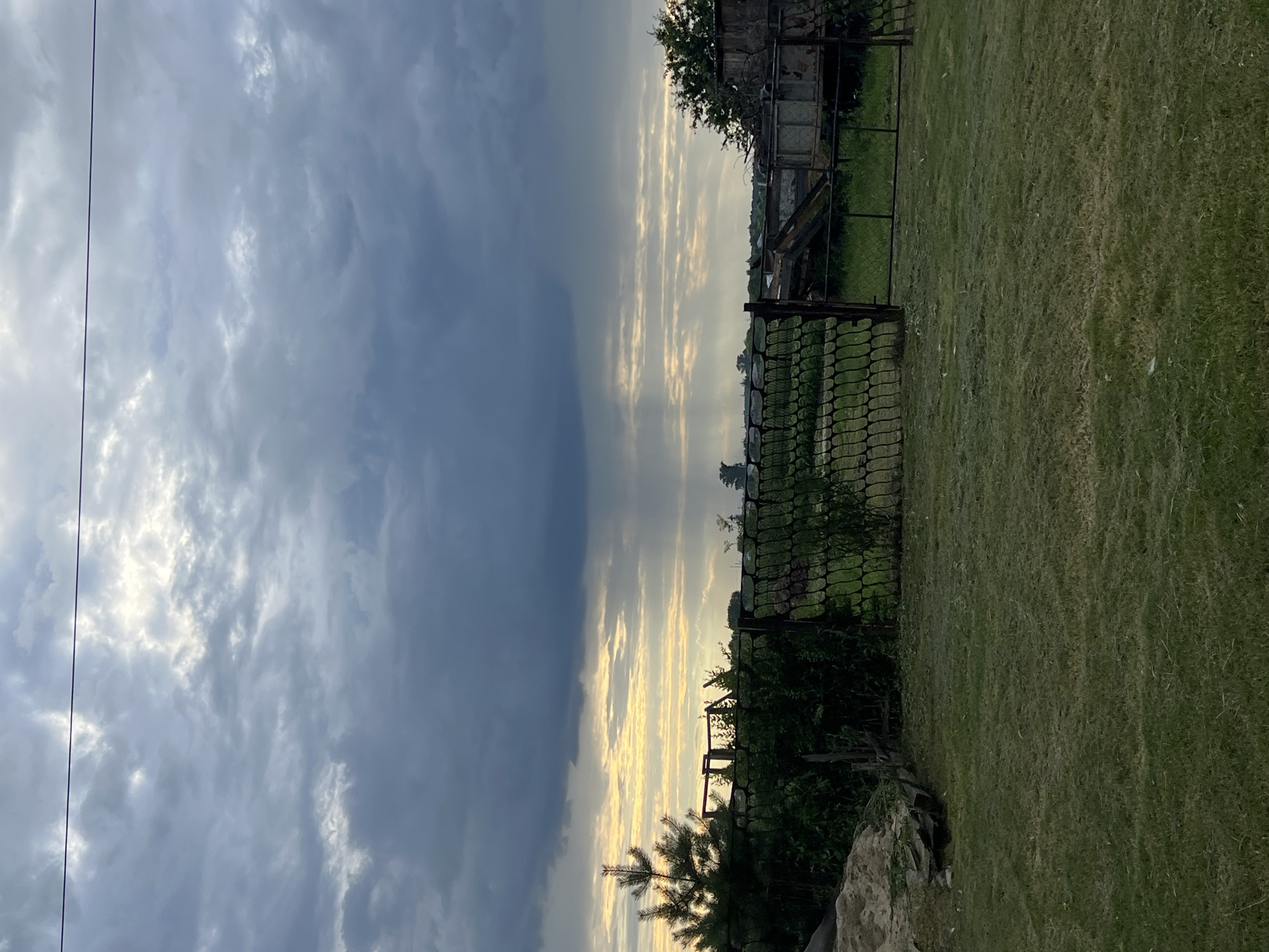 The image shows an outdoor scene with a wire fence in the foreground. Beyond the fence, there is a grassy area with some trees and shrubs. In the background, there are buildings and a large, cloudy sky with rays of sunlight breaking through. The scene appears to be a rural area, possibly in the city of Pivkino, Russia, given the context provided. The lighting suggests it is either early morning or late evening. There are no people visible in the image.