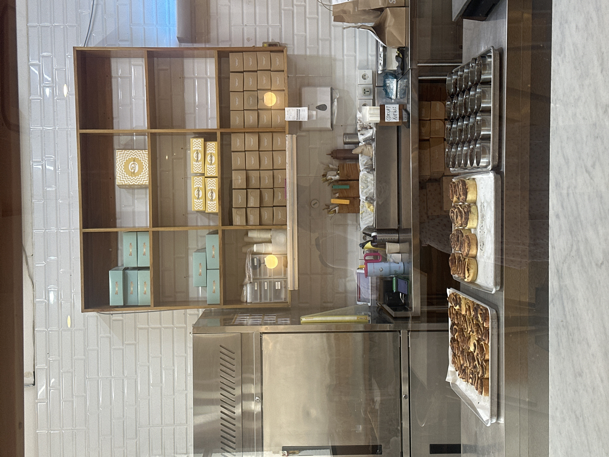 The image depicts a bakery counter in North Kuta, Indonesia, viewed through a glass window. In the foreground, two trays of cinnamon rolls are displayed on a stainless steel counter. To the right of the cinnamon rolls, a tray of round metal molds filled with what appears to be raw dough is visible. Behind these, a shelf unit with several compartments is stocked with various boxes, some teal and others brown, with labels. A commercial refrigerator with a stainless steel exterior is positioned to the left. The background features a white tiled wall and a counter area with cups, containers, and other bakery supplies. A sign on the counter reads "COKLAT NEWS!". The scene suggests a retail bakery preparing or displaying its products.