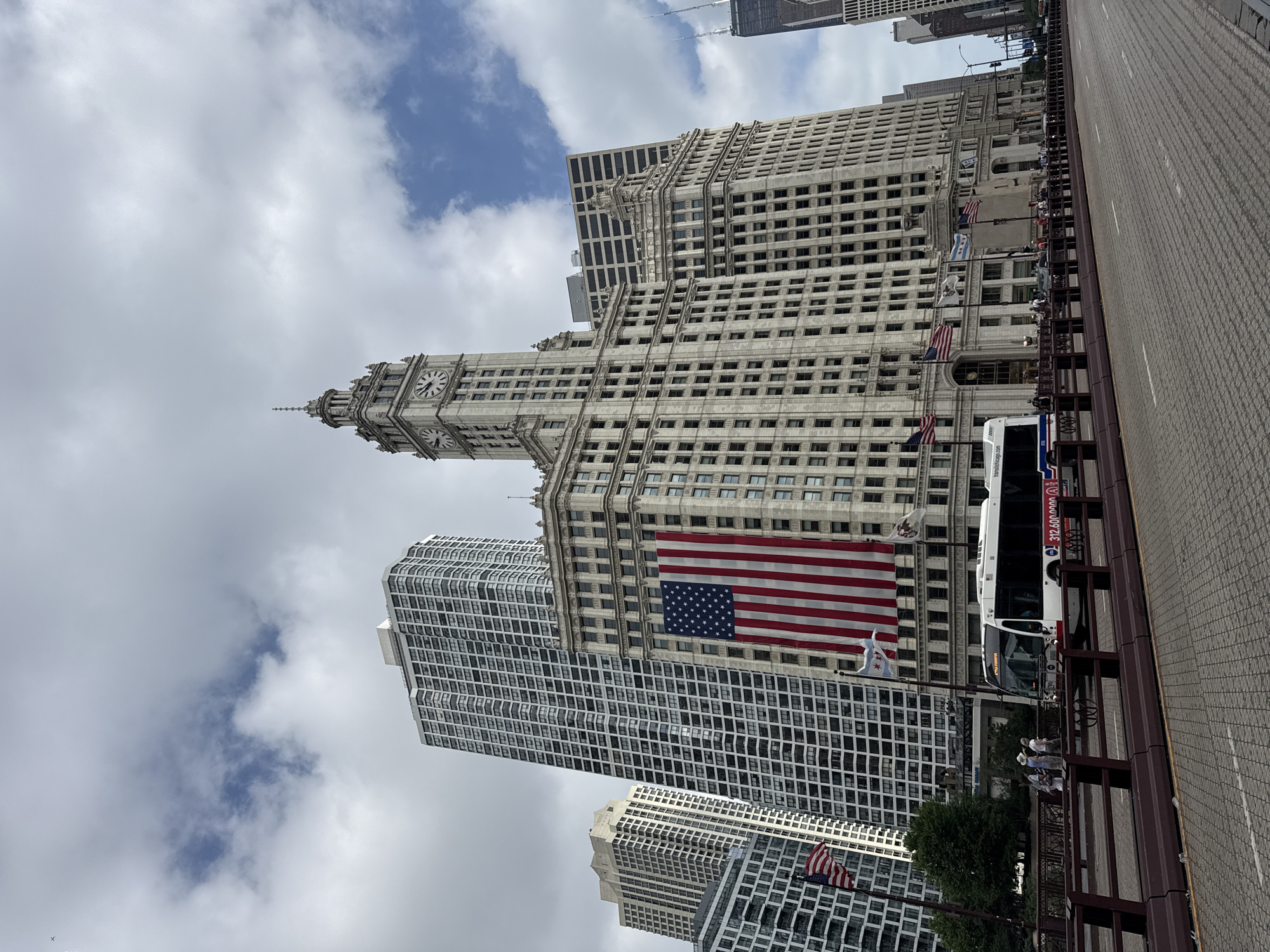 This is a ground-level, eye-level photograph of the Wrigley Building in Chicago, Illinois, United States of America, captured on a cloudy day. The iconic white terra cotta building with its prominent clock tower is the central focus, draped with a large American flag. To the left of the Wrigley Building, the tall, slender white residential skyscraper at 1000 N Lake Shore Plaza and other modern high-rises are visible. In the foreground, a street and a pedestrian walkway with a railing are present. A white bus with "transitchicago.com" on its side is traveling on the road, and several people are walking on the sidewalk. Several other flags are also visible, including the Chicago city flag. The sky is partially obscured by clouds, with patches of blue showing through.