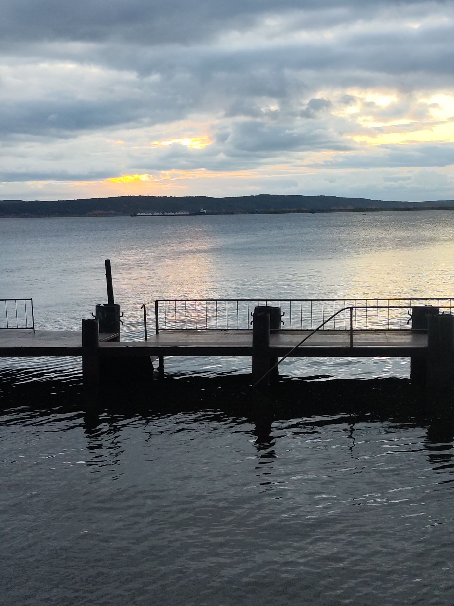The image depicts a pier extending into a calm body of water under a cloudy sky with patches of sunlight breaking through. The pier, made of wood, has metal railings and dark posts. Reflections of the pier and the sky are visible on the water's surface. In the distance, a treed shoreline forms the horizon, with a barge visible on the water. The overall scene is serene and captures a moment at dusk. This scene is located at the Kama River embankment in Chaikovsky, Russia.