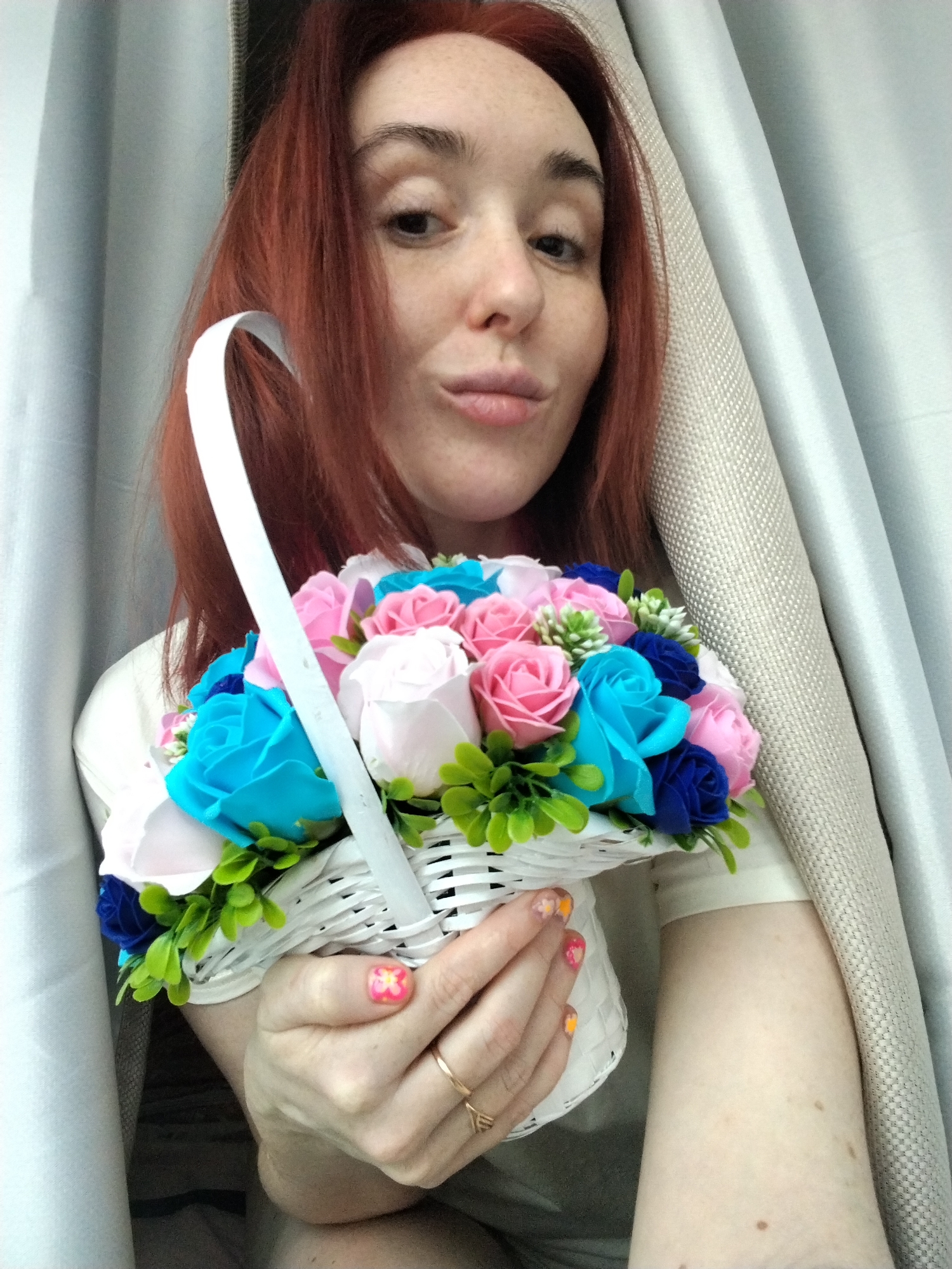 An adult female is depicted indoors in Izhevsk, Russia, shown in a medium close-up, chest-up shot. She has reddish-brown hair and a fair complexion with visible freckles across her nose and cheeks. Her left eye is fully visible, while her right eye is partially obscured by her hair. Her lips are pursed, and she looks directly at the camera. She is wearing a light-colored, short-sleeved garment. Both hands are holding a white woven basket filled with an arrangement of artificial, rose-shaped flowers in shades of bright pink, light pink, light blue (cyan), and dark blue, accented with green foliage and small white buds. Her fingernails are painted with a multi-colored floral design. A plain gold-colored ring is present on the ring finger of her left hand. The background features draped curtains, with a light grey or off-white curtain on the left and a beige or light brown textured curtain on the right.