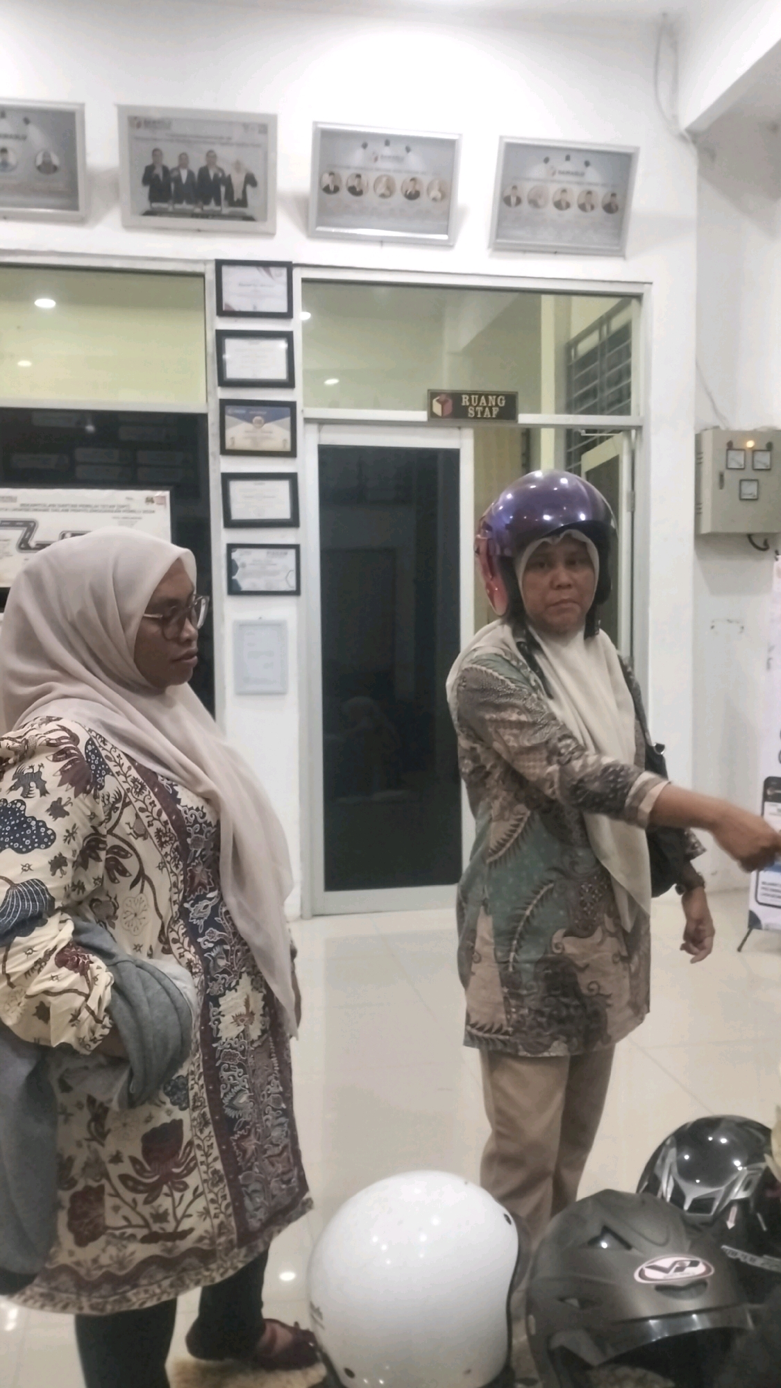 The image captures an indoor scene, likely an office or a public building, in Lhokseumawe, Indonesia. Two women are the primary subjects. The woman on the left wears a beige hijab and a patterned dress with brown, white, and blue motifs. She has glasses and is looking to her left. The woman on the right is wearing a purple helmet and a patterned top with green, brown, and white designs, along with beige trousers. She is looking towards the right side of the frame and her right arm is extended, with her hand pointed.
The environment features a wall with framed pictures and certificates. One sign on the wall reads "RUANG STAF" which translates to "Staff Room" in Indonesian. There are also helmets visible in the foreground, suggesting the presence of motorcycles or a mode of transportation requiring helmets. The lighting appears to be artificial.