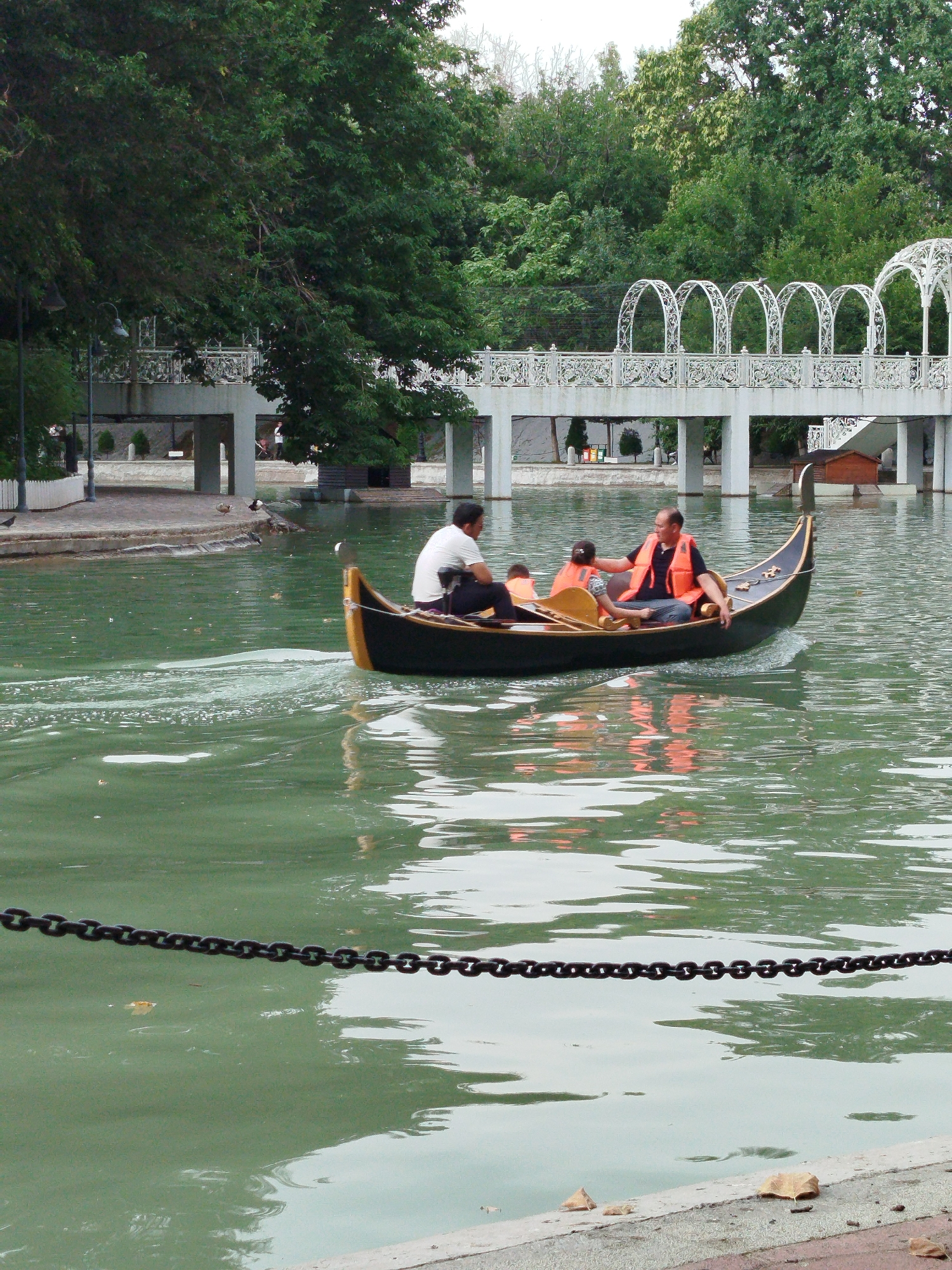 The image depicts a gondola boat with four occupants navigating a tranquil, green body of water. The boat, a dark color with yellow accents, leaves a visible wake. Two adults and two children are visible, all wearing bright orange life vests, suggesting a recreational activity. One adult in the front appears to be guiding the boat. The backdrop features lush green trees and an arched pedestrian bridge with white railings and decorative white arches. The water surface reflects the surrounding greenery and the sky. In the foreground, a dark chain is stretched across the water, a few fallen leaves are scattered on the embankment, and a paved walkway is visible. The scene is suggestive of a park or recreational area. This specific scene is located in the "Anhor Lokomotiv" park in Tashkent, Uzbekistan, known for its picturesque canal and recreational facilities.