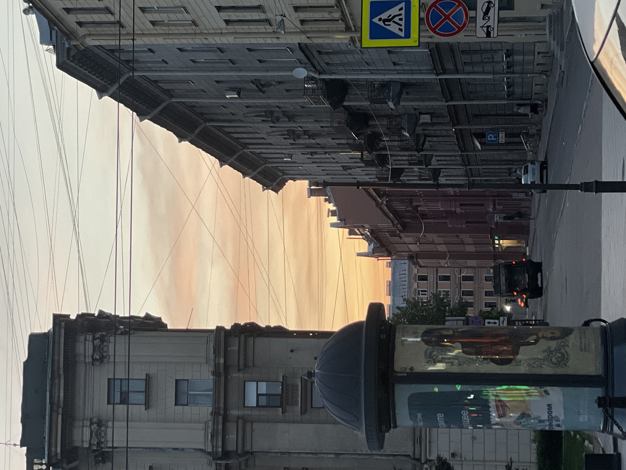 The image captures a street view in Saint Petersburg, Russia, at dusk or dawn. The sky exhibits a gradient of colors, ranging from pale yellows and oranges near the horizon to muted grays higher up. Buildings line both sides of the street, showcasing classical architecture with detailed facades. Several overhead power lines are crisscrossing the sky.
On the right side of the frame, a street sign is visible, and further down the street, there is a black vehicle with its headlights on. On the left, a light post stands with advertisements. The scene is set on an urban street, capturing a moment of daily life in the city.