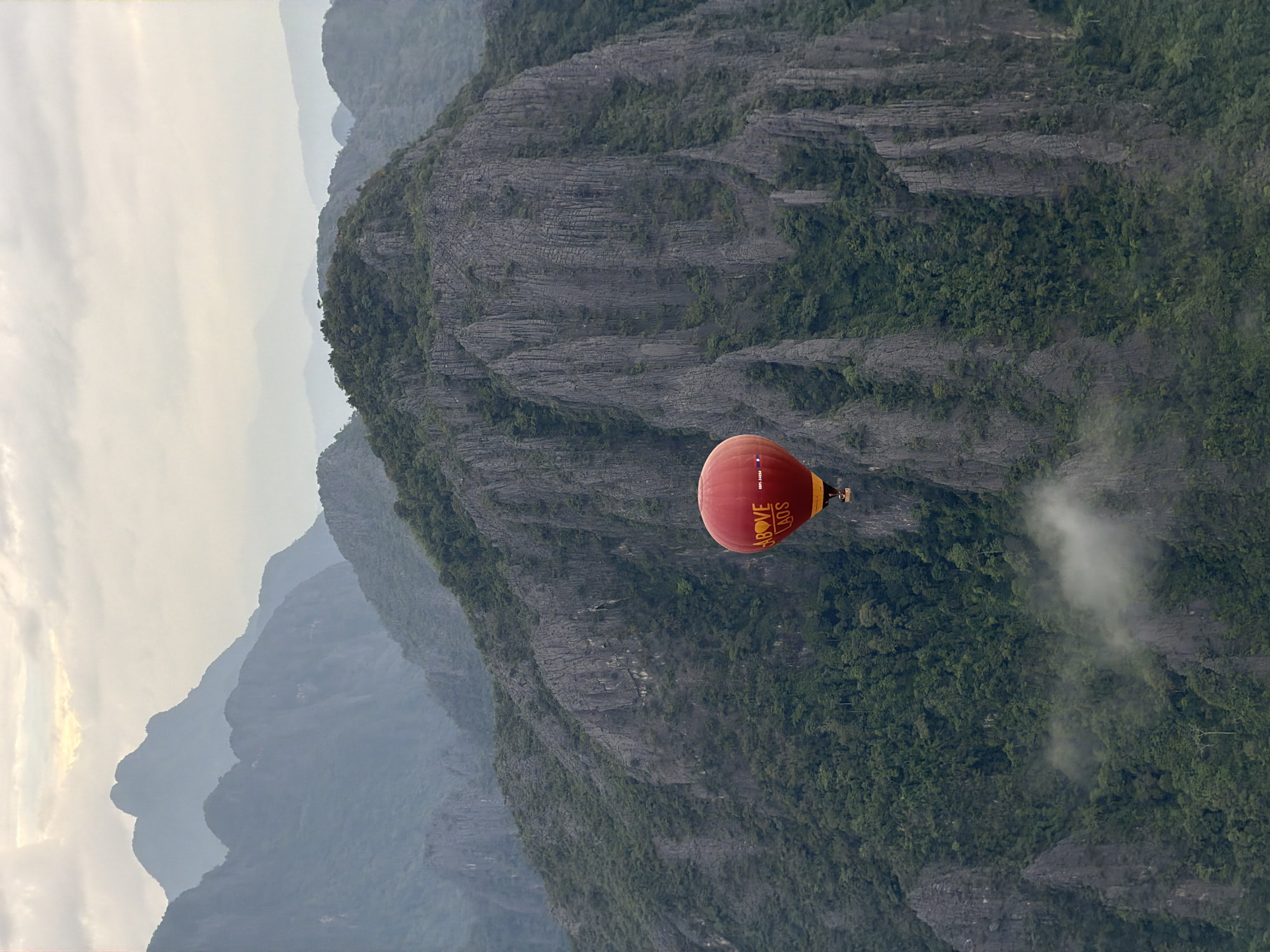 A red hot air balloon with the words "Above Laos" written on it in yellow lettering is captured in flight against a backdrop of lush green mountains and a hazy sky. The balloon is positioned in the lower right quadrant of the image. The mountains are steep and rugged, with dense vegetation covering their slopes. Wisps of cloud are visible amongst the peaks, adding a sense of depth and atmosphere to the scene. The sky is overcast, with diffused light suggesting it may be early morning or late afternoon. The image appears to be taken from another elevated position, likely a nearby mountain or another hot air balloon. The overall impression is one of serene natural beauty and adventure, characteristic of Vang Vieng, Laos, a popular destination for hot air ballooning due to its dramatic karst landscape.
