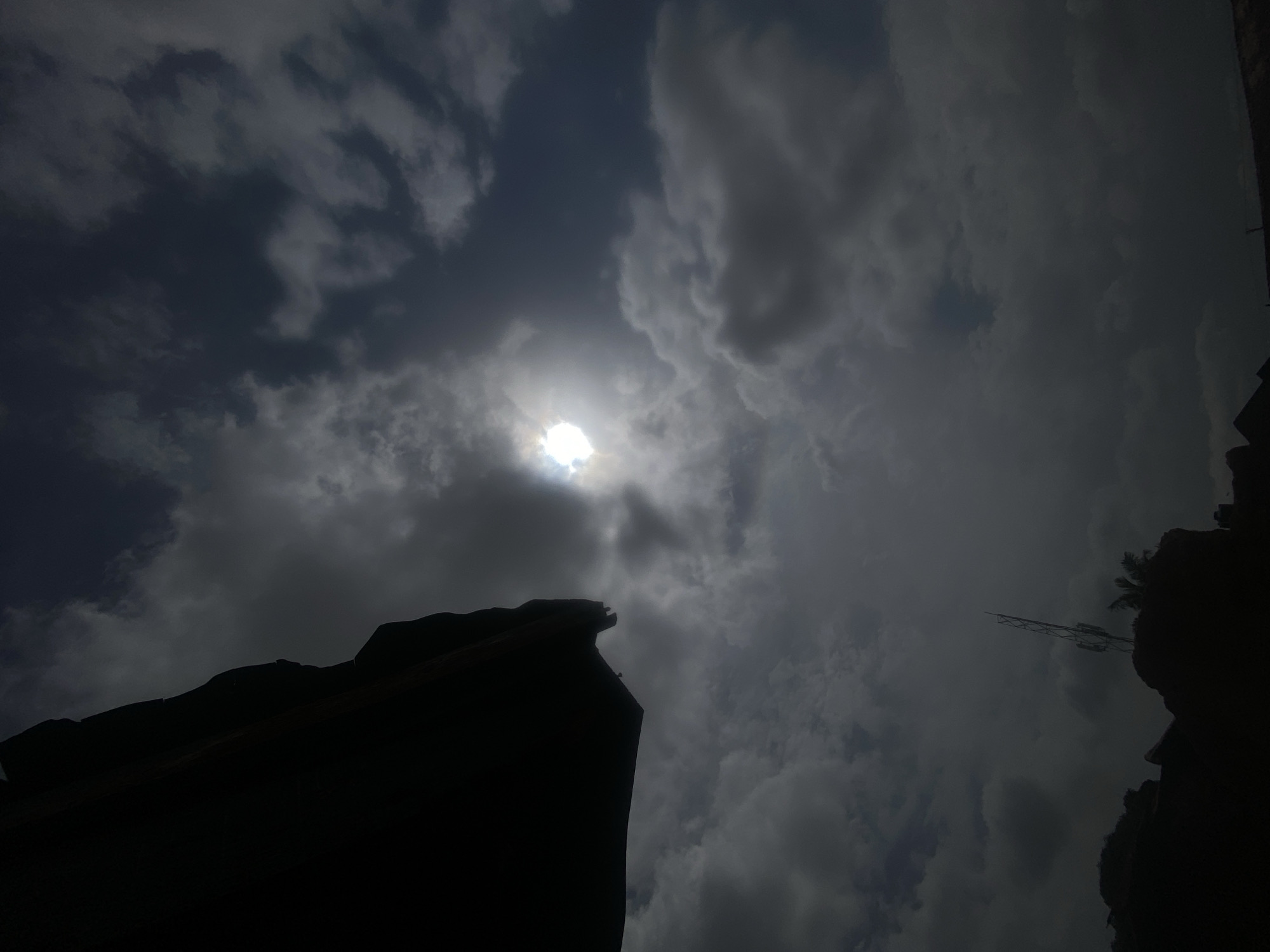 The image shows a low-angle view towards the sky. The sun is visible through the clouds, creating a diffused light. Silhouetted in the foreground are the outlines of structures. In the right corner, there is a tower-like structure visible. It is daytime. It is not possible to ascertain the location as Barden Yana from this image.