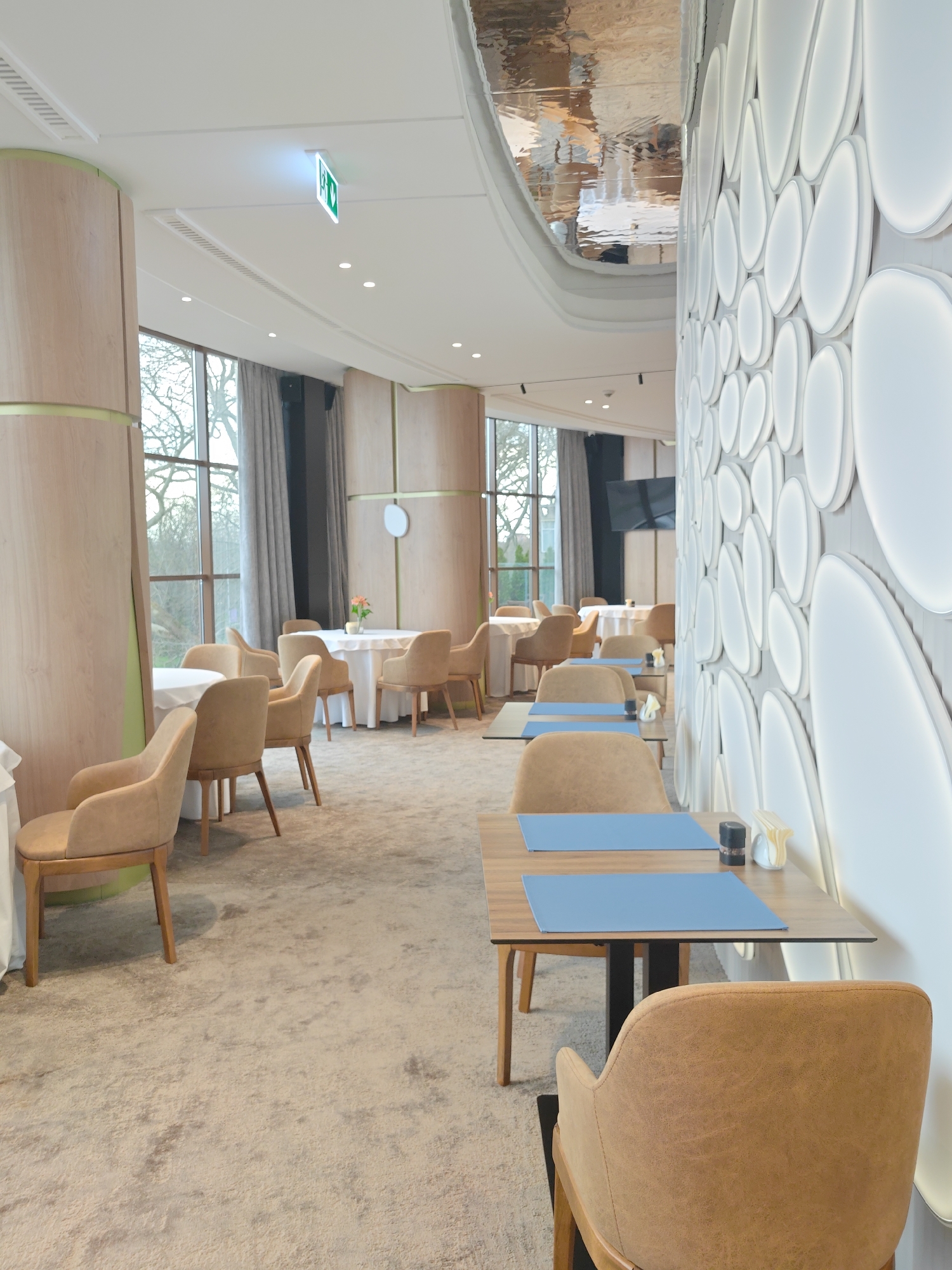 The image shows the interior of a restaurant in Varna, Bulgaria. The dining area features tables with light-brown wooden surfaces and blue placemats. The chairs are light brown with a leather-like material and wooden frames. The flooring is carpeted in a beige color with a mottled texture. There are large windows covered by grey curtains allowing the view to an outside area with vegetation. The wall is decorated with white, egg-shaped, illuminated elements. The ceiling is mostly white, and there is a mirrored section. An emergency exit sign is visible. Some tables are covered with white tablecloths and decorated with flowers.