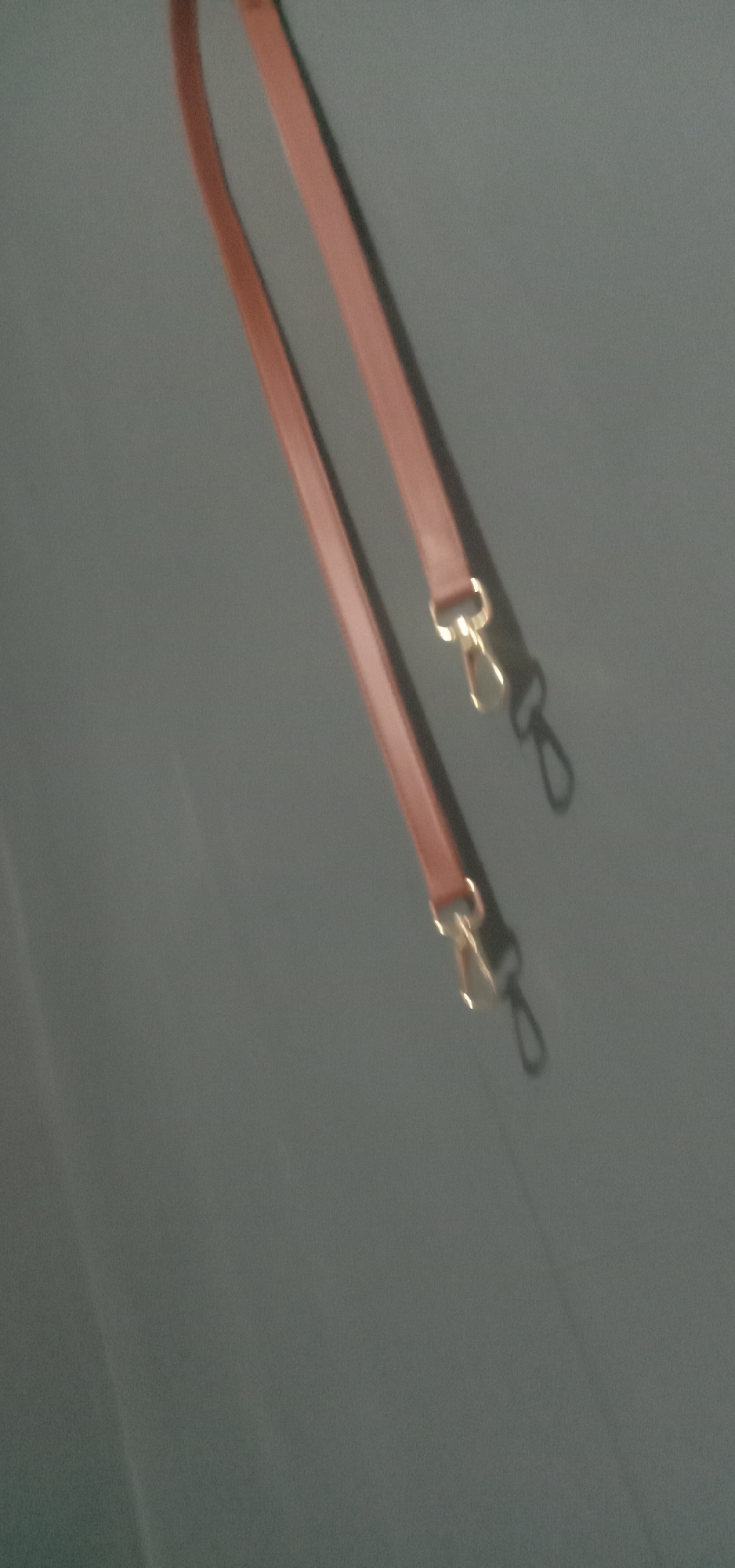 The image displays a pair of brown leather straps with gold-colored clasps against a dark gray background. The straps are positioned diagonally, with the top strap appearing slightly above and to the right of the bottom strap. Both straps have a rectangular end with a metal loop through which the clasps are attached. The clasps are designed to swivel and appear to be of a standard hook-and-eye type. The background is smooth and featureless, suggesting a flat surface or fabric. There are no people, animals, or other objects visible in the image, nor is there any indication of a specific location in Kendari, Indonesia. The lighting suggests an overhead light source, casting subtle shadows from the straps onto the background. The image is taken from a slightly elevated angle, looking down at the straps.