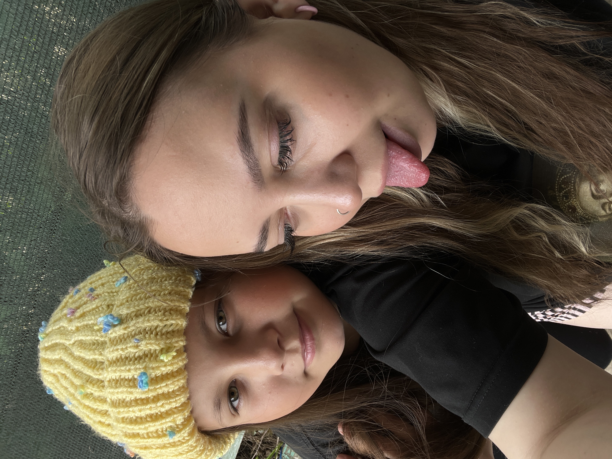 Two females are posing closely together for a photo. The female at the top of the frame is sticking her tongue out. She has brown hair with blonde highlights, fair skin, a nose ring, and long eyelashes. The female at the bottom of the frame is wearing a yellow knit hat with blue and yellow embellishments. She has fair skin, brown hair, and brown eyes. Both females are wearing black tops. The background appears to be an outdoor setting, possibly a garden or backyard, with a green mesh-like surface visible in the top-left corner. There is no information to confirm or deny the location of the scene in Novopokrovskaya, Russia.