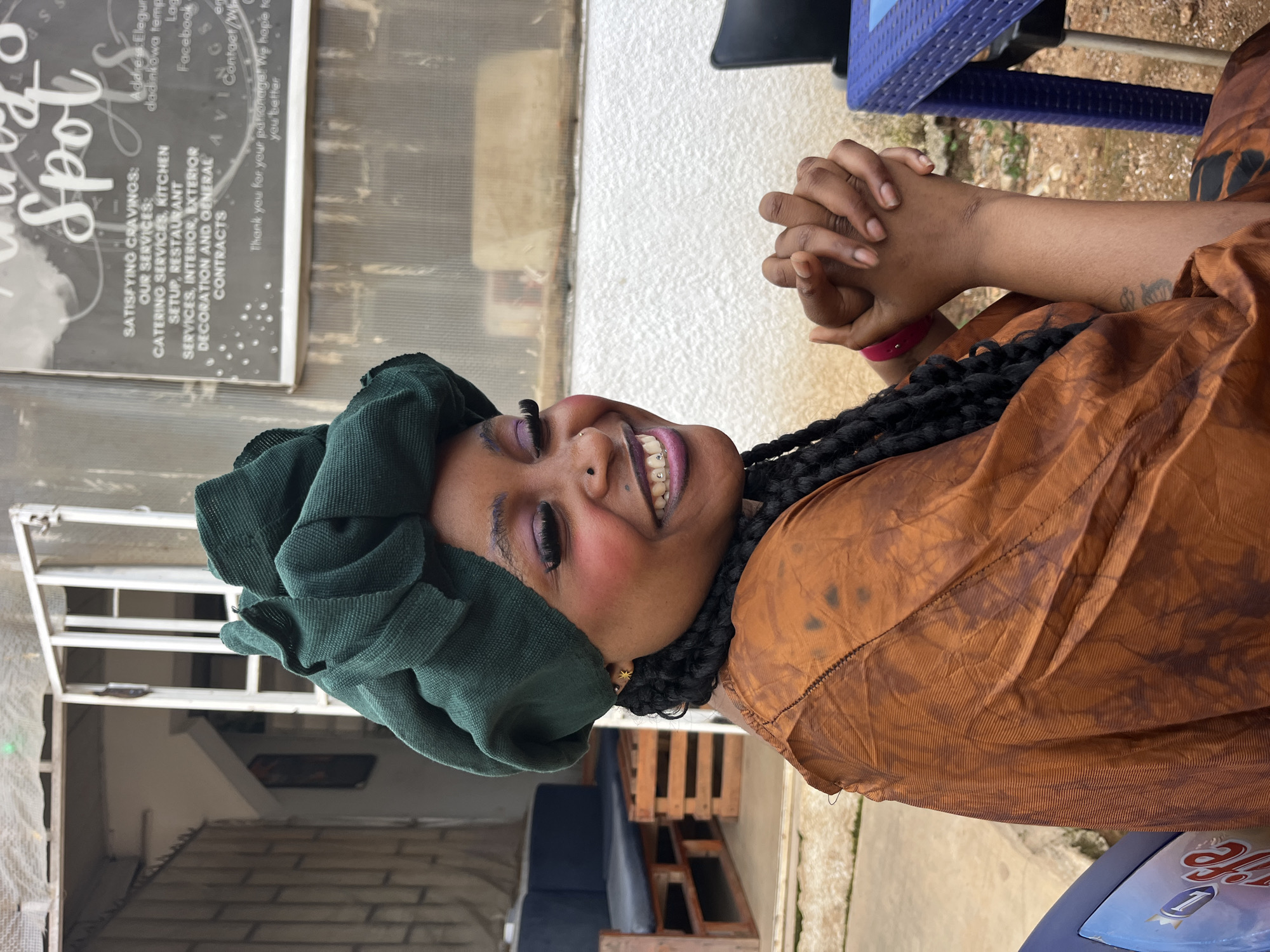 A woman with dark skin, a green headwrap, and a brown dress is sitting outdoors, smiling at the camera. She has long eyelashes, purple eyeshadow, and a nose ring. Her hands are clasped in her lap. In the background, there is a building with a sign that reads "Tasty Spot." There are also some pallets and a blue table visible. The setting appears to be a casual outdoor dining area.