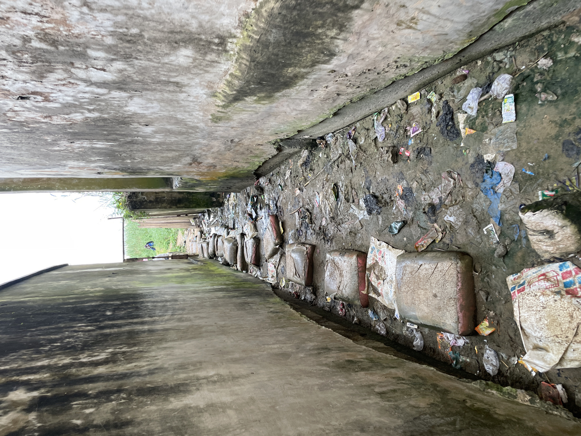 A narrow, muddy pathway is flanked by two weathered concrete walls with dark streaks and patches of green moss. The ground is covered in a mix of dirt, stagnant water, and a significant amount of scattered trash, including plastic bags, wrappers, and bottles. A line of large, rectangular objects, possibly sacks or bags, are placed as stepping stones along the left side of the path, leading towards a brighter opening at the end. In the distance, at the end of the alley, a person wearing a blue shirt and dark pants is visible, standing in a grassy area. The overall scene depicts an unsanitary and neglected urban environment.