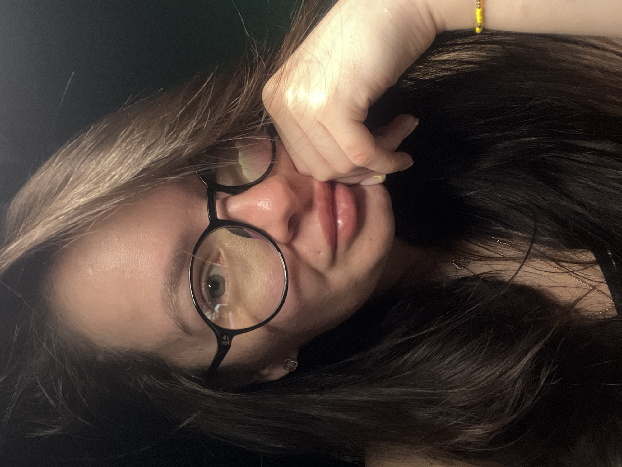 A close-up, vertically-oriented selfie captures a young woman with light skin, dark hair streaked with lighter shades, and wearing round black eyeglasses. Her right hand is positioned near her face, with fingers gently touching her cheek and lips. The background is predominantly dark, creating contrast with her face and hair. A small gold chain necklace is visible around her neck. There's a yellow beaded bracelet on her wrist. A small earring can be seen. The image shows reflections in her glasses. The location in the city of Kazan, Russia, cannot be determined from the given image.