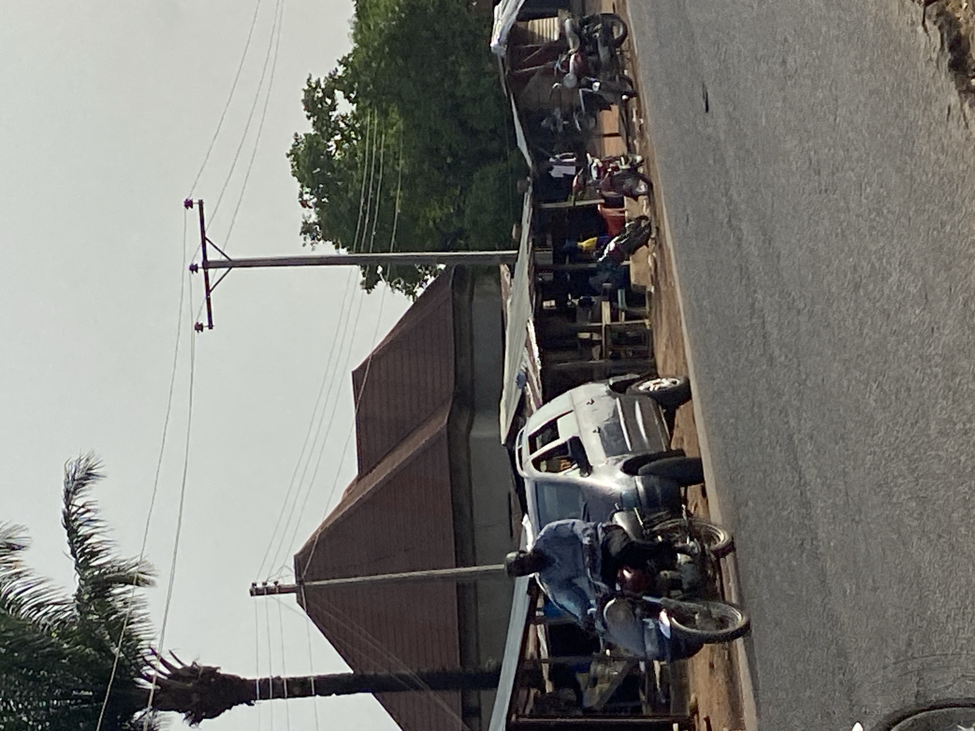 This media file captures a daytime street scene in Mararaba, Nigeria, depicting a busy roadside environment. The primary focus is along a paved road with various activities and structures.
In the foreground, a person is seen riding a motorcycle, heading away from the viewer on the right side of the road. Further down, another white van or minibus is parked on the side of the road, and several other motorcycles are visible, some parked, suggesting a common mode of transport and commerce in the area.
Along the left side of the road, a series of low-rise buildings or stalls with corrugated metal roofs line the street, indicating commercial activity. A tall utility pole with multiple power lines extends upwards from this side, with its wires stretching across the frame. Another utility pole is visible further back. Lush green trees, including the top of a palm tree with its distinct fronds and trunk, are interspersed between and behind the buildings, providing natural elements to the urban landscape.
The sky is overcast and appears bright, suggesting a daytime setting, likely late morning or afternoon, without direct harsh sunlight. There is no discernible text visible in the image. The overall impression is that of a bustling, everyday street scene in a Nigerian town.