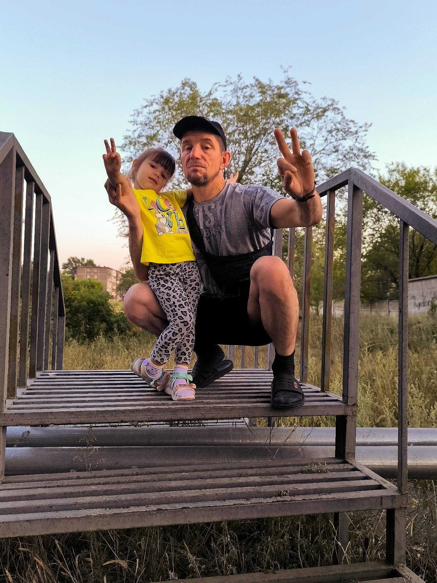 A man and a young girl are posing for a photograph on a metal staircase outdoors. The man, wearing a black baseball cap, a grey t-shirt, and black shorts, is crouched down. The girl, in a yellow t-shirt and patterned leggings, stands in front of him. Both are holding up two fingers in a peace sign. The background shows dry grass, trees, and buildings, suggesting an urban or suburban environment. The image's metadata does not contain information about the specific location within Qarağandı city administration, Kazakhstan.