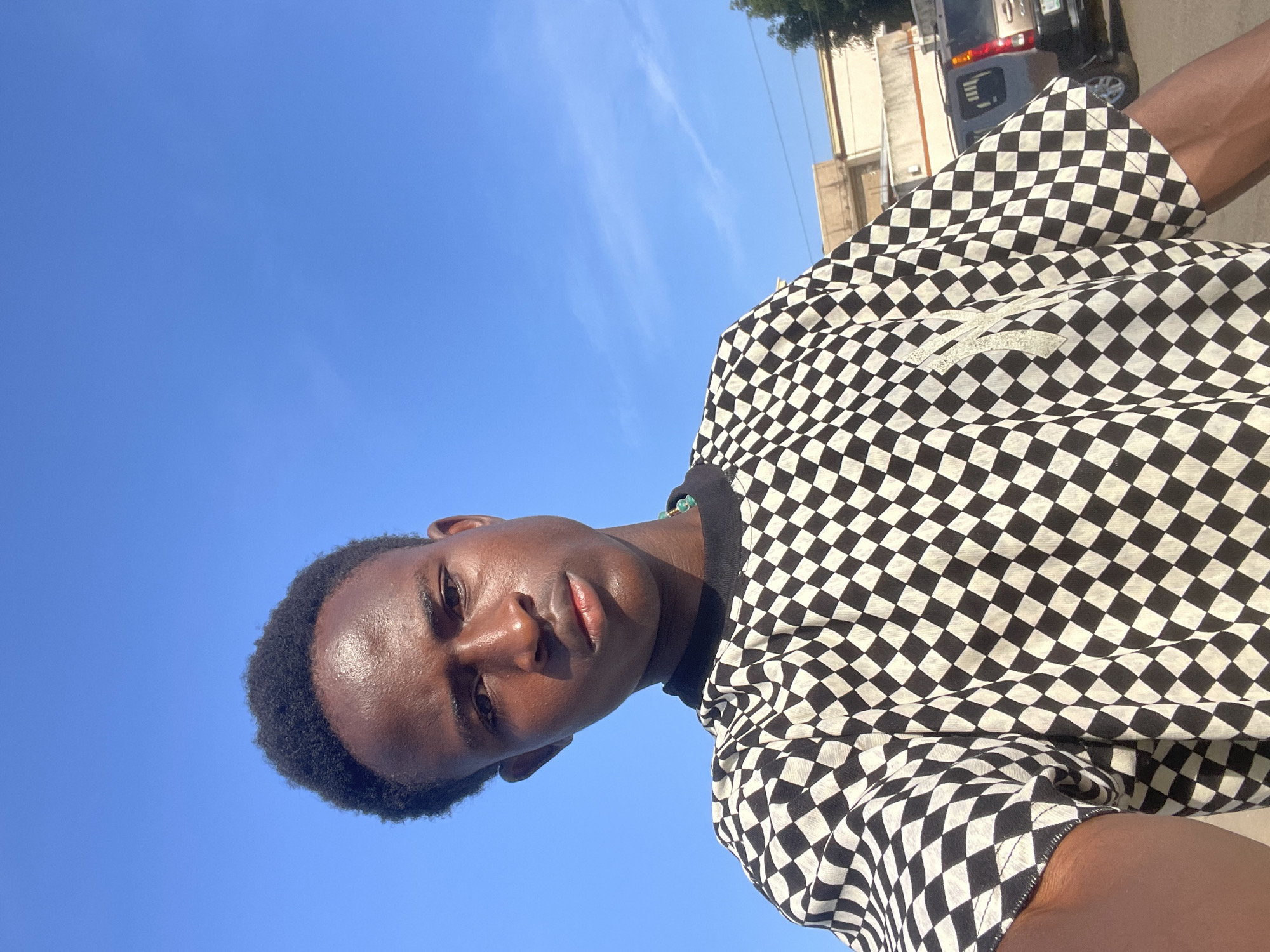 A young man with dark skin and short, curly hair is pictured from the chest up, looking directly at the camera. He is wearing a black and white checkered t-shirt with a small, light-colored logo on the left chest. The background is a clear blue sky, with a hint of clouds. In the distance, there are buildings and a parked vehicle, suggesting an outdoor urban setting. The image was taken in Jos, Nigeria.