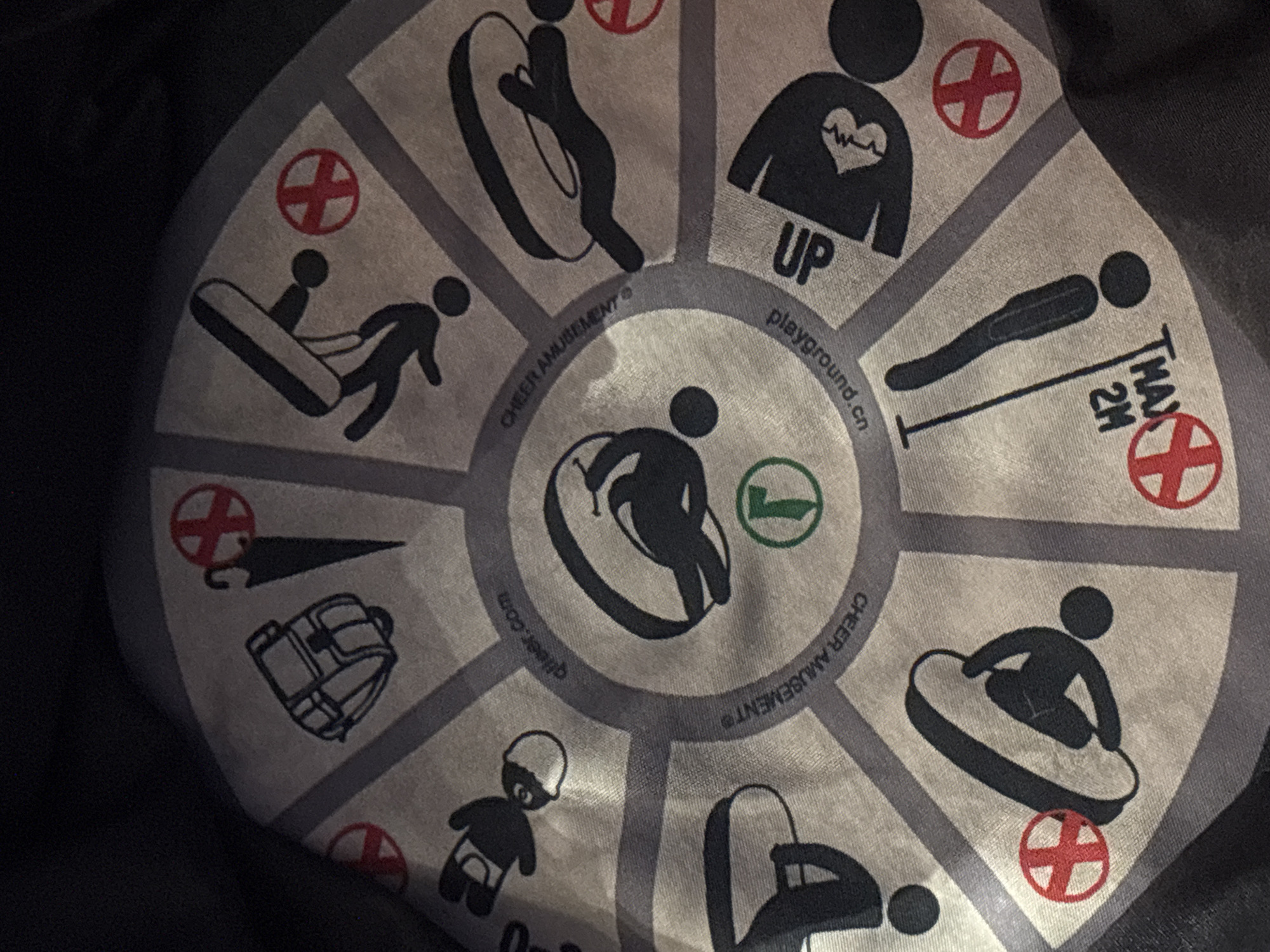 The image shows a close-up view of a circular textile pattern featuring various icons arranged radially. The pattern appears to be a safety or instructional guide, possibly for a recreational activity or product.
Specific icons visible include:
* A backpack.
* An umbrella with a red cross symbol overlay.
* A figure in a prone position, possibly performing a stunt.
* A figure holding a toy.
* A figure on a steering wheel.
* A figure with a heart monitor line.
* A figure falling.
* An exclamation mark with text that appears to say "MAX 2H" and "UP".
There are also red circular symbols with a white cross, and a green circular symbol with a stylized "T" or "F". Text elements include "qiqeer.com", "CHEER AMUSEMENT®", and "playground.cn".
The textile is depicted against a dark, out-of-focus background, suggesting it is either a garment, a tarp, or a similar fabric item. The lighting is dim, and the image is taken from a slightly elevated angle, looking down onto the patterned surface.
There is no information in the image to suggest a specific location in Dubai, United Arab Emirates. The text elements present are likely company or website names associated with the product or service depicted by the icons.
